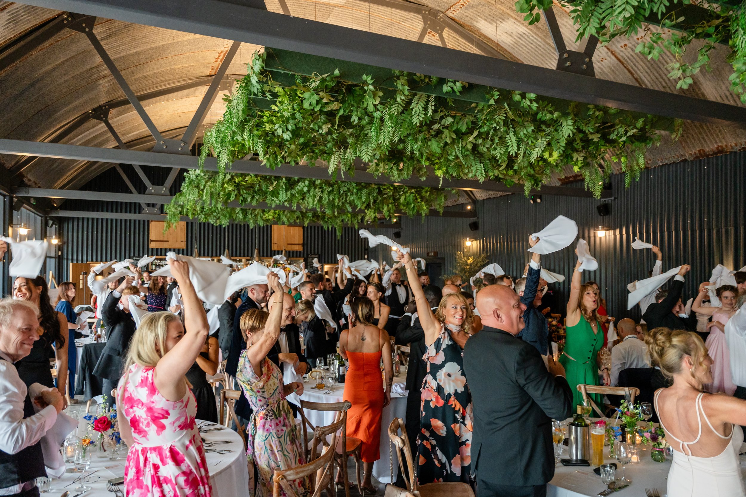 People celebrating at a large indoor event, waving napkins in the air, with a green hanging plant installation and industrial-style decor.