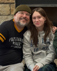 A man and a young woman sitting together, smiling at the camera. The man has a beard and glasses, wearing a dark beanie and black shirt. The young woman has long brown hair, wearing a gray hoodie and sitting close to him.
