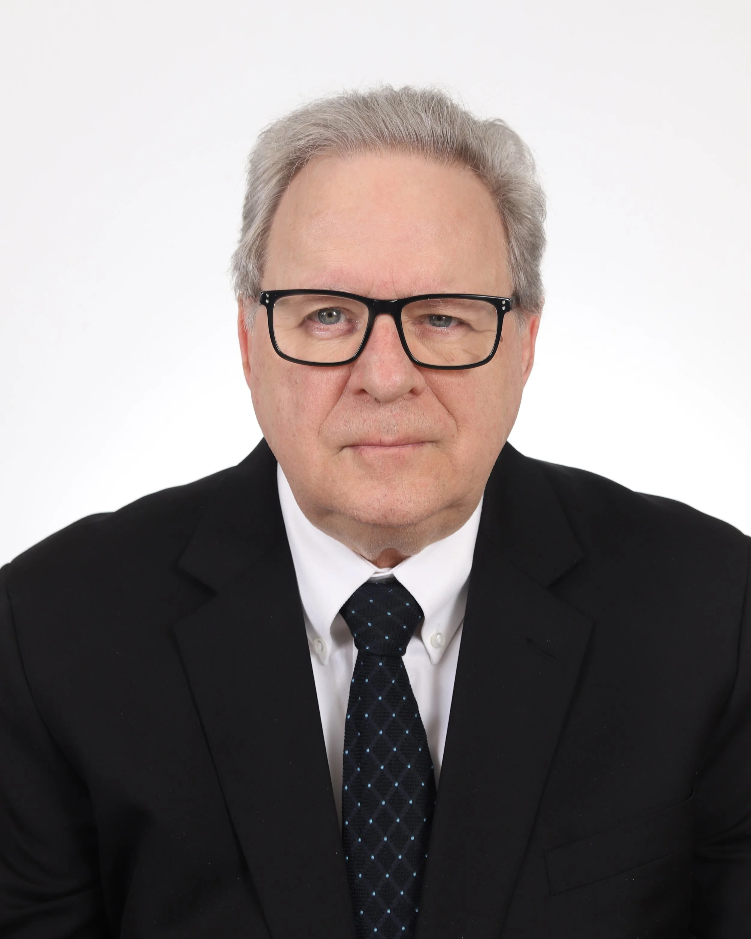 A middle-aged man with gray hair, wearing glasses, a dark suit, a light blue shirt, and a patterned tie, looking directly at the camera with a serious expression.