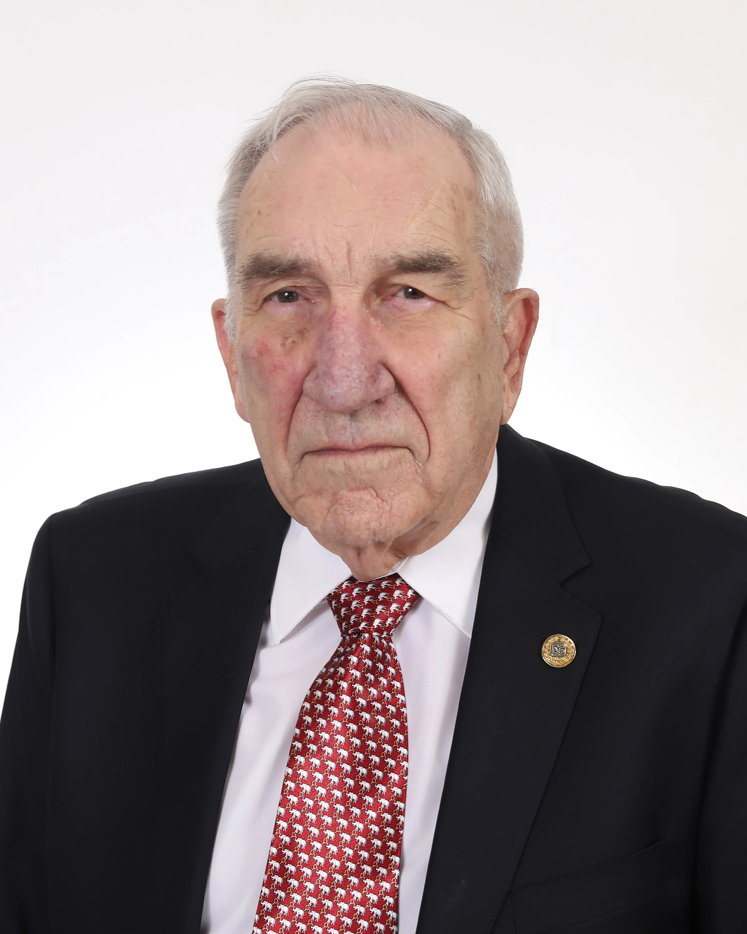 Close-up of an elderly man wearing a suit and tie with a serious expression, dark background.