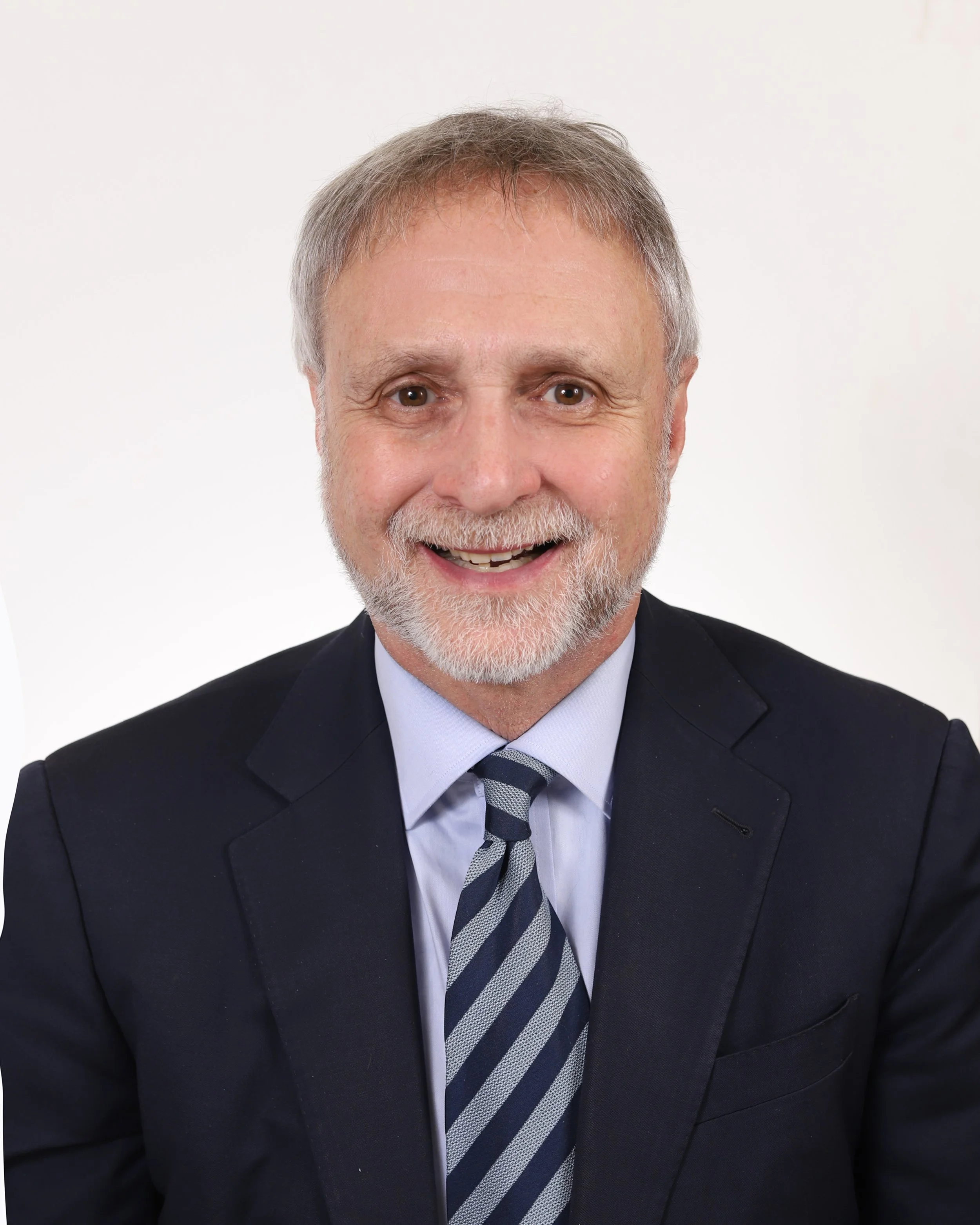 A professional headshot of a middle-aged man with gray hair, a beard, wearing a dark suit, white shirt, and blue tie, smiling at the camera.