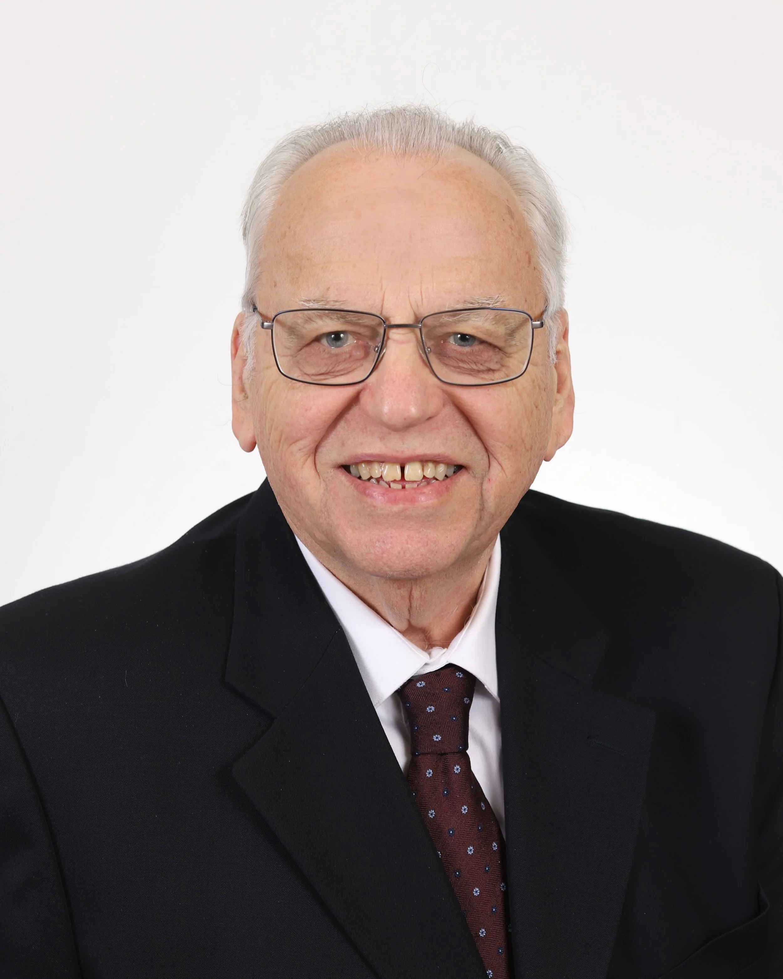 A smiling elderly man with white hair and glasses, dressed in a dark suit, white shirt, and patterned tie, against a black background.