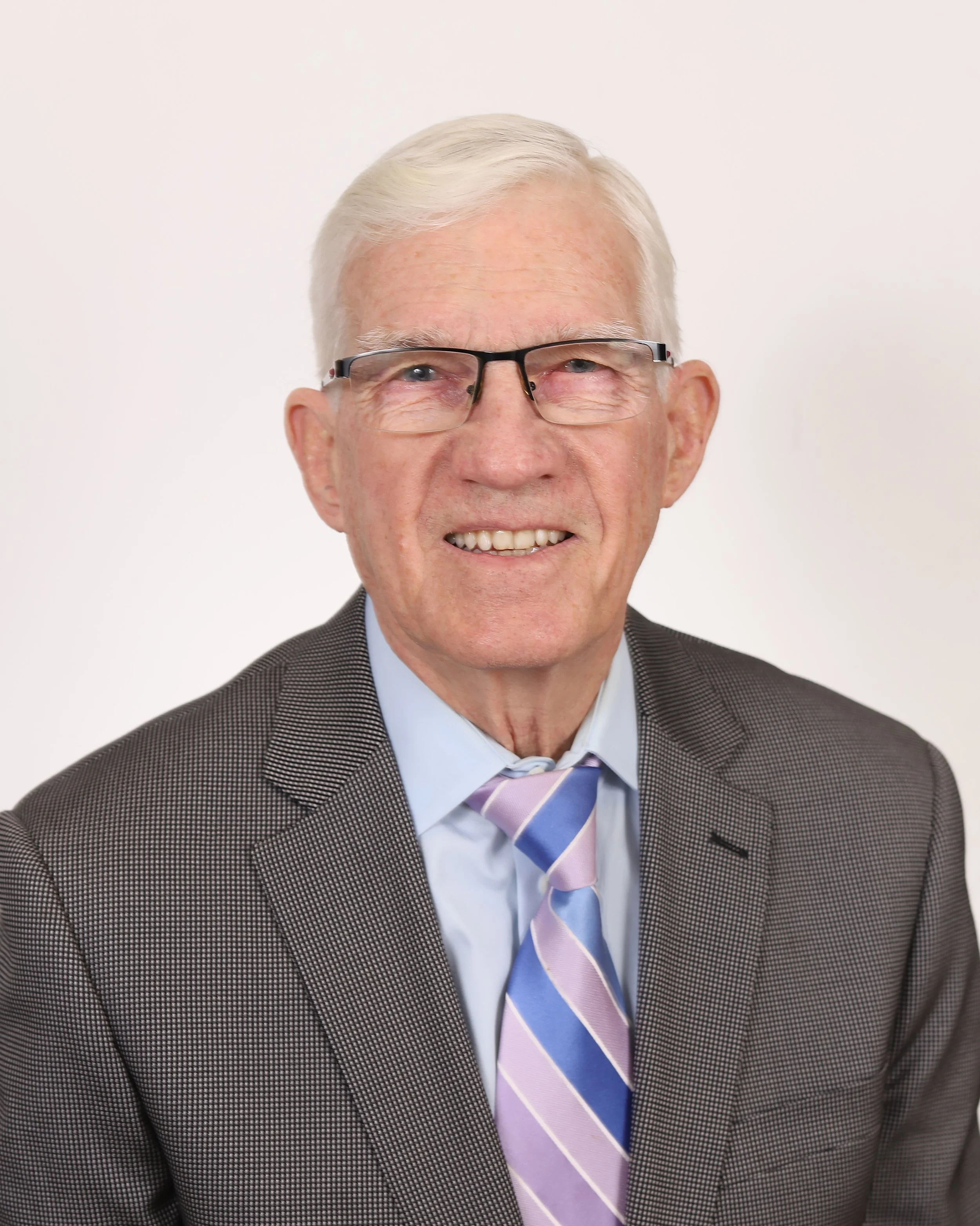 An older man with white hair, wearing glasses, a light-colored, checkered dress shirt, and a patterned tie, smiling against a black background.