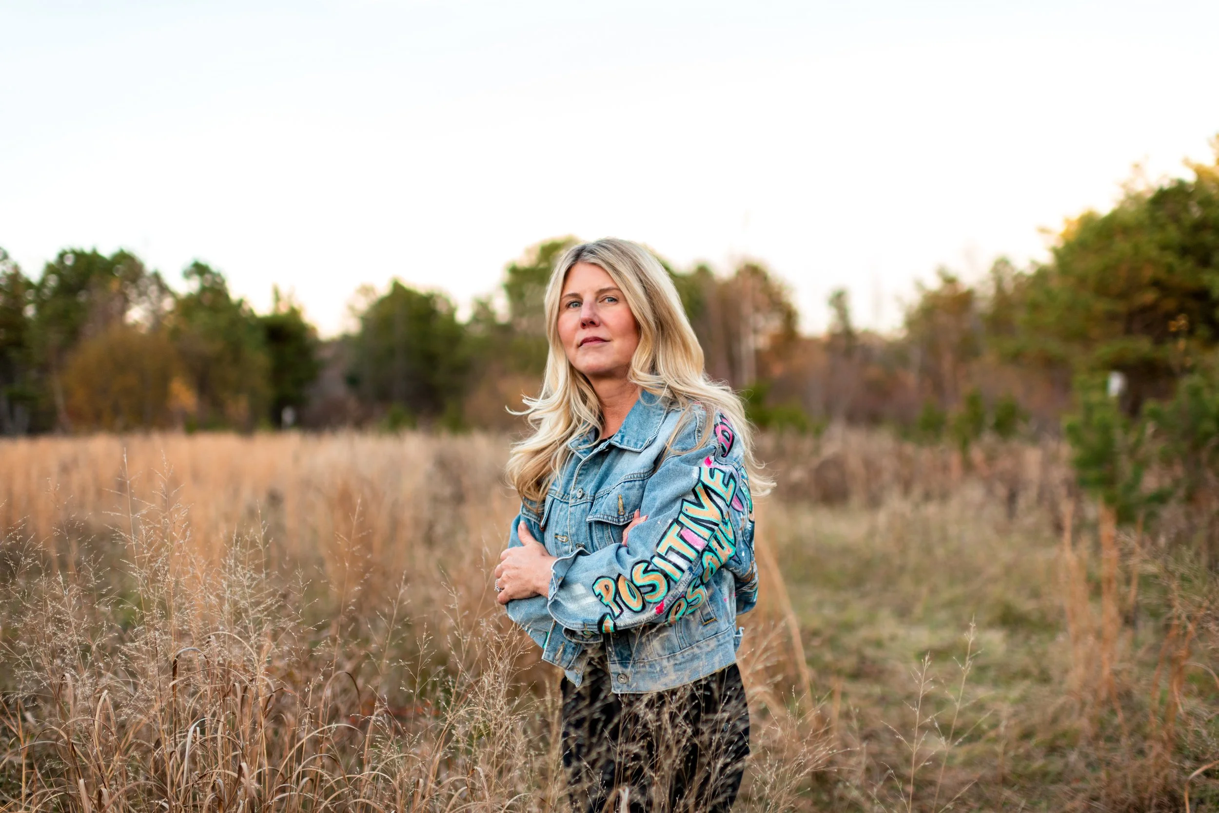 A woman standing outdoors in a field with tall dry grass, wearing a blue denim jacket with colorful text on the sleeve, and looking at the camera. The background shows trees and a clear sky.