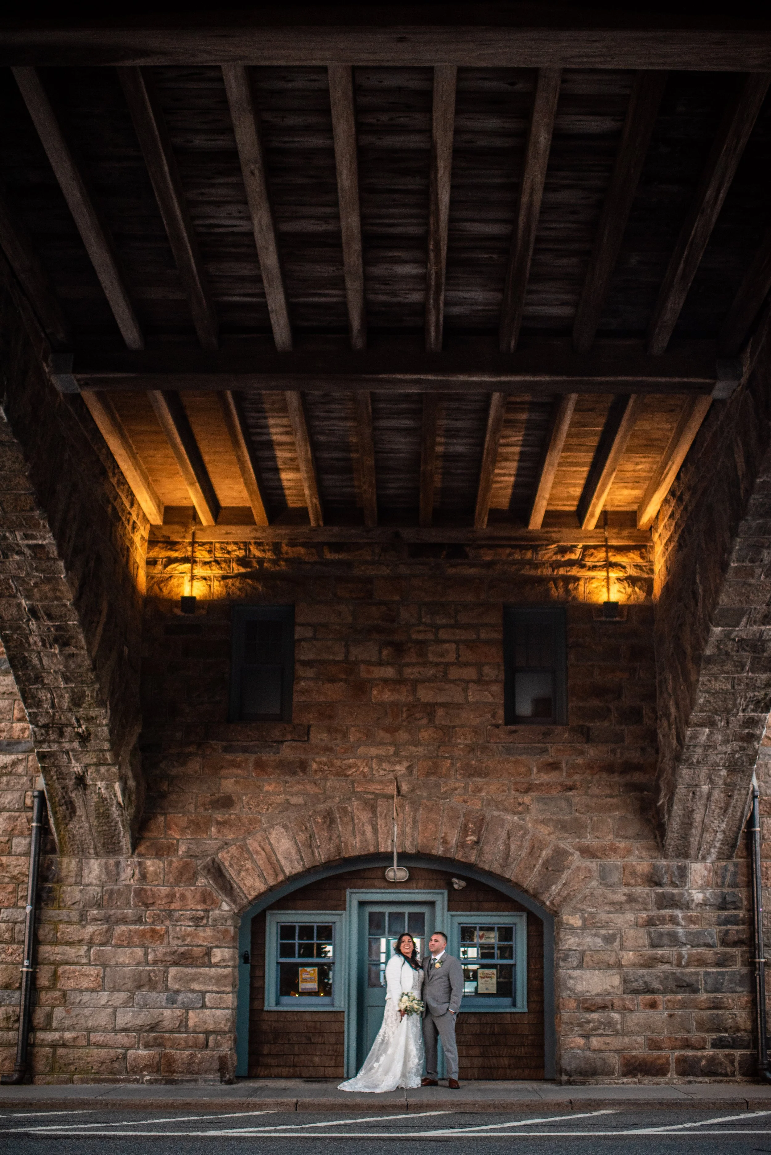 A bride and groom in wedding attire standing under an archway of an old brick building at night.