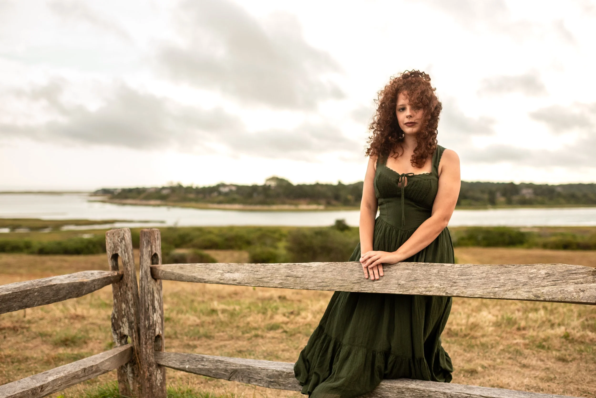 A woman with curly red hair wearing a dark green dress outdoors, standing behind a wooden fence, with water and cloudy sky in the background.