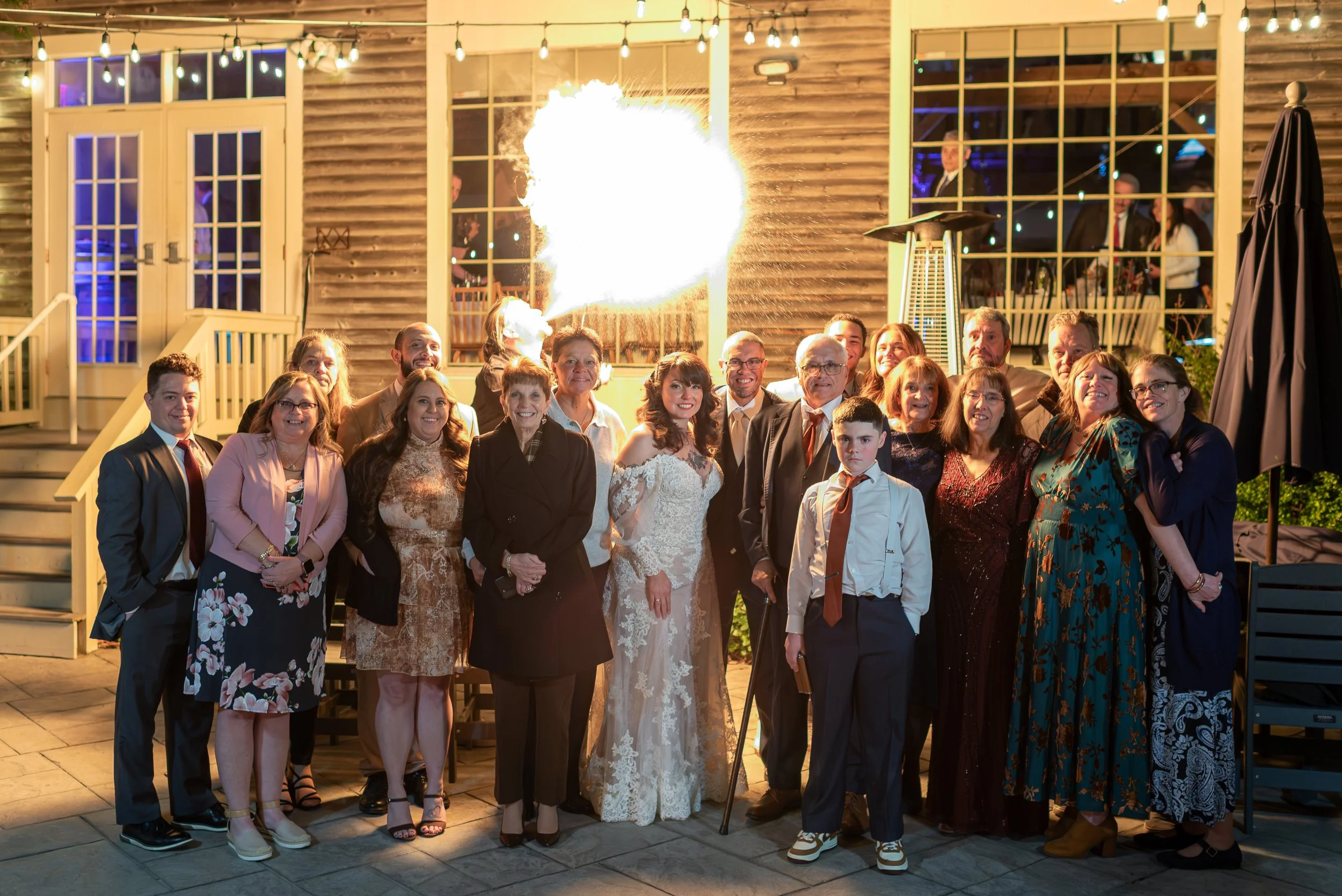 Group of people celebrating a wedding outdoors at night with string lights, a large fire, and a rustic building in the background.