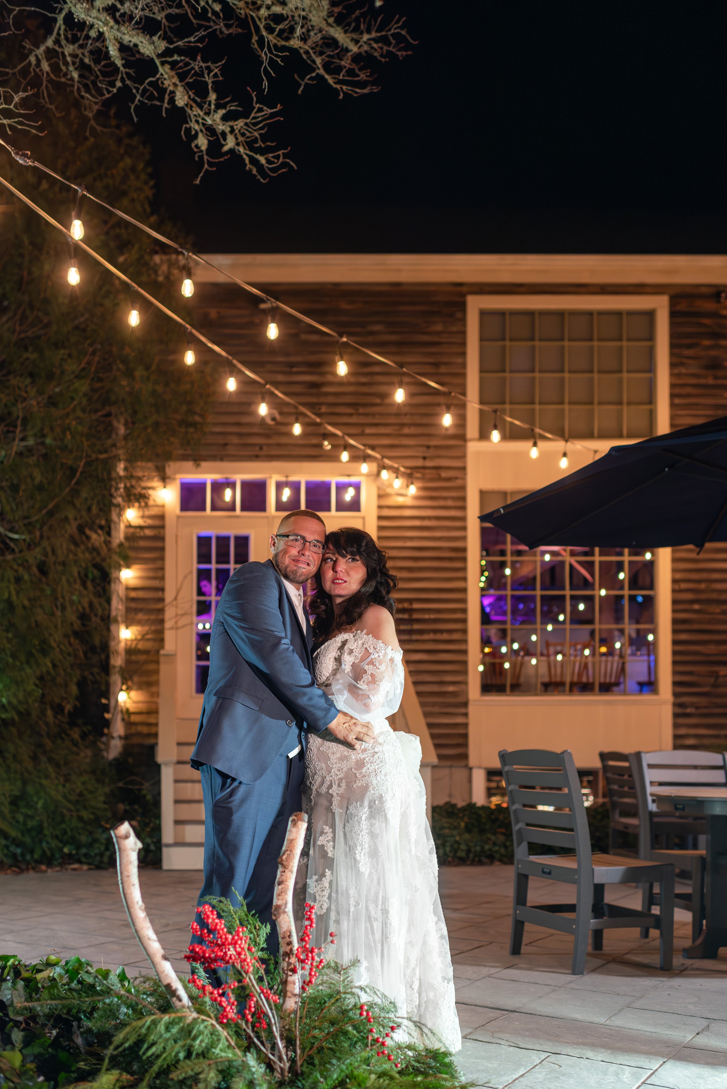 A newlywed couple embracing outdoors at night under string lights, with a rustic wooden building and decorated patio behind them.