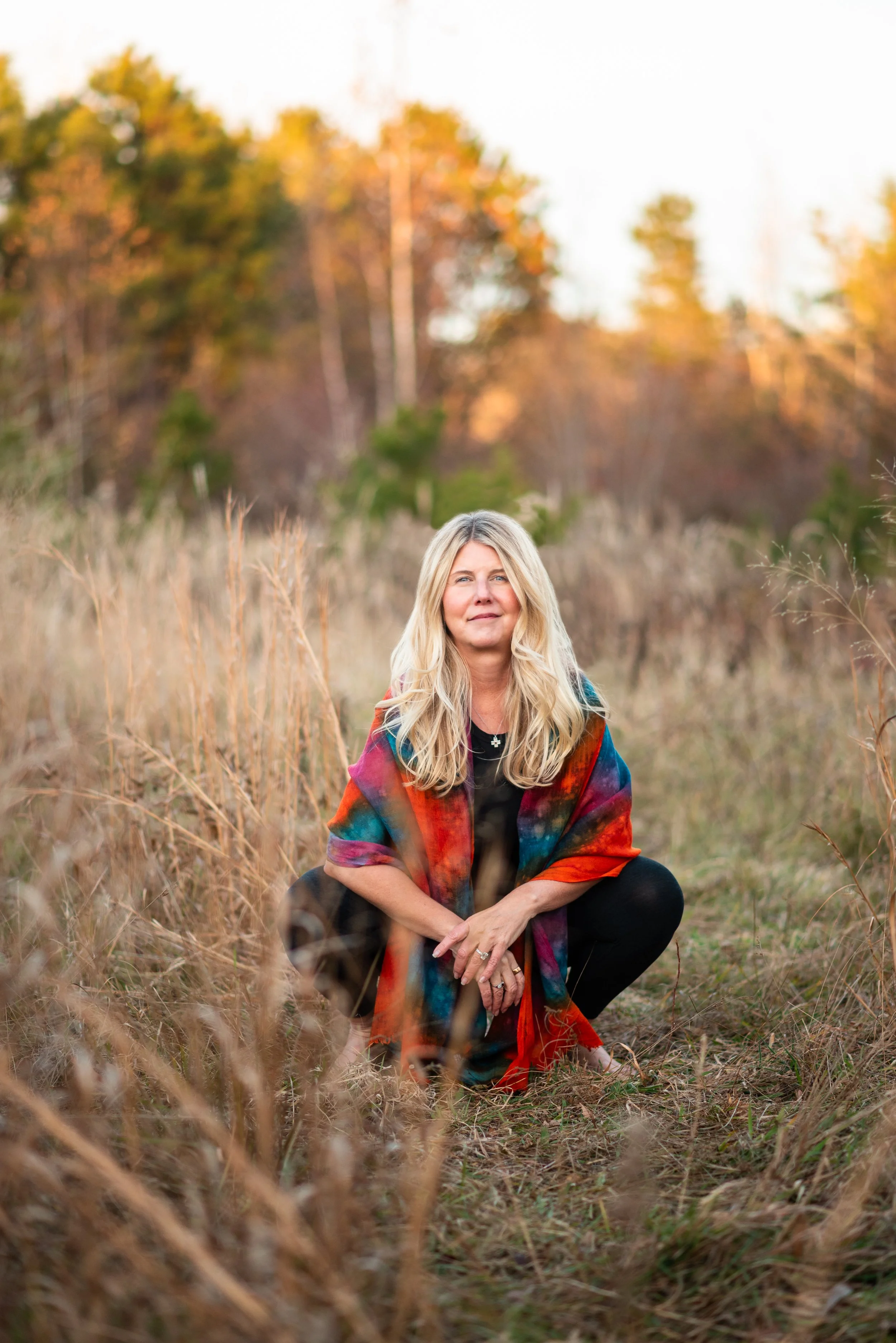 A woman with blonde hair crouches in a field of tall, dry grass during sunset, wearing a colorful shawl and black clothing, with a blurred forest in the background.