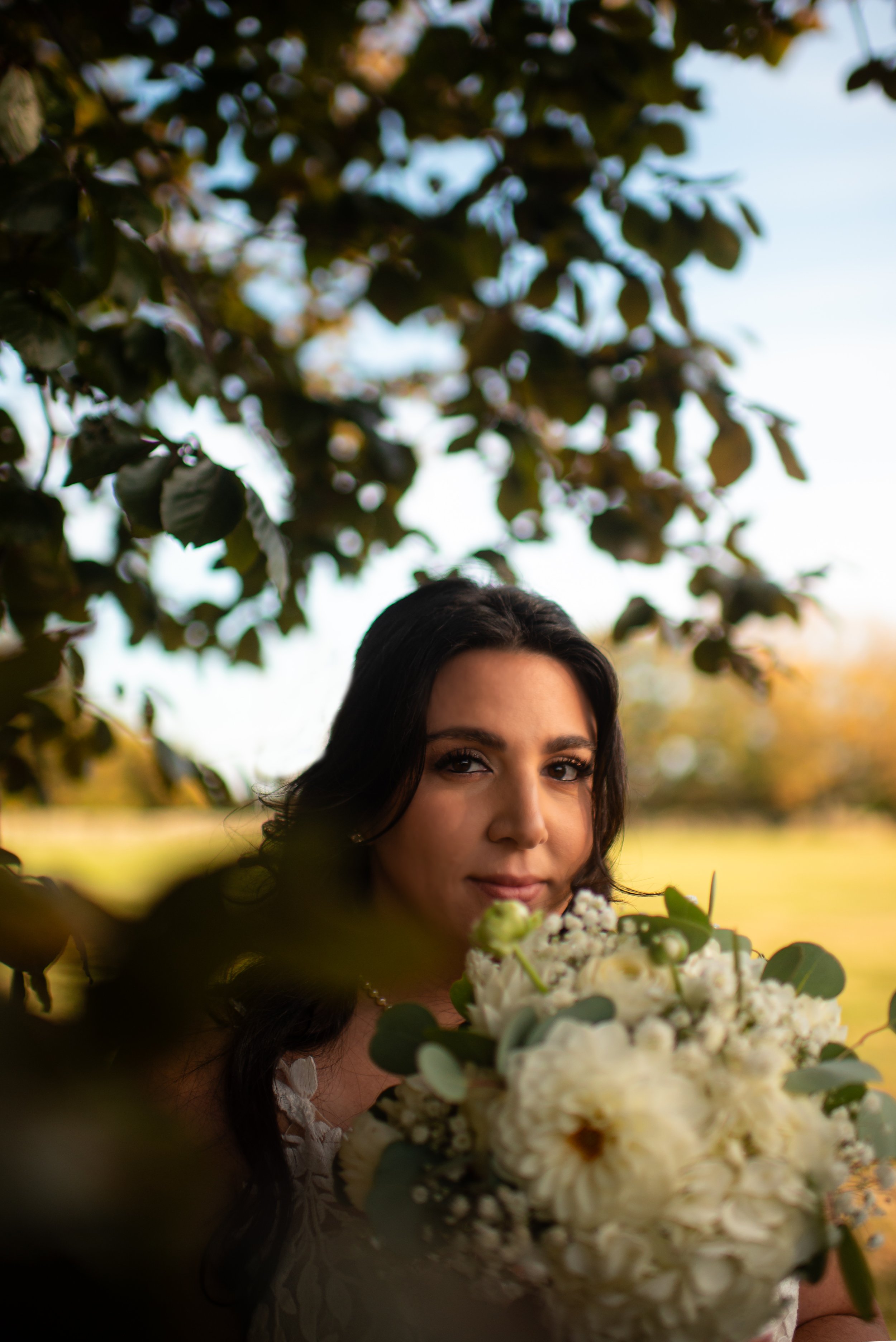 A woman holding a bouquet of white flowers outdoors, framed by tree branches and leaves.