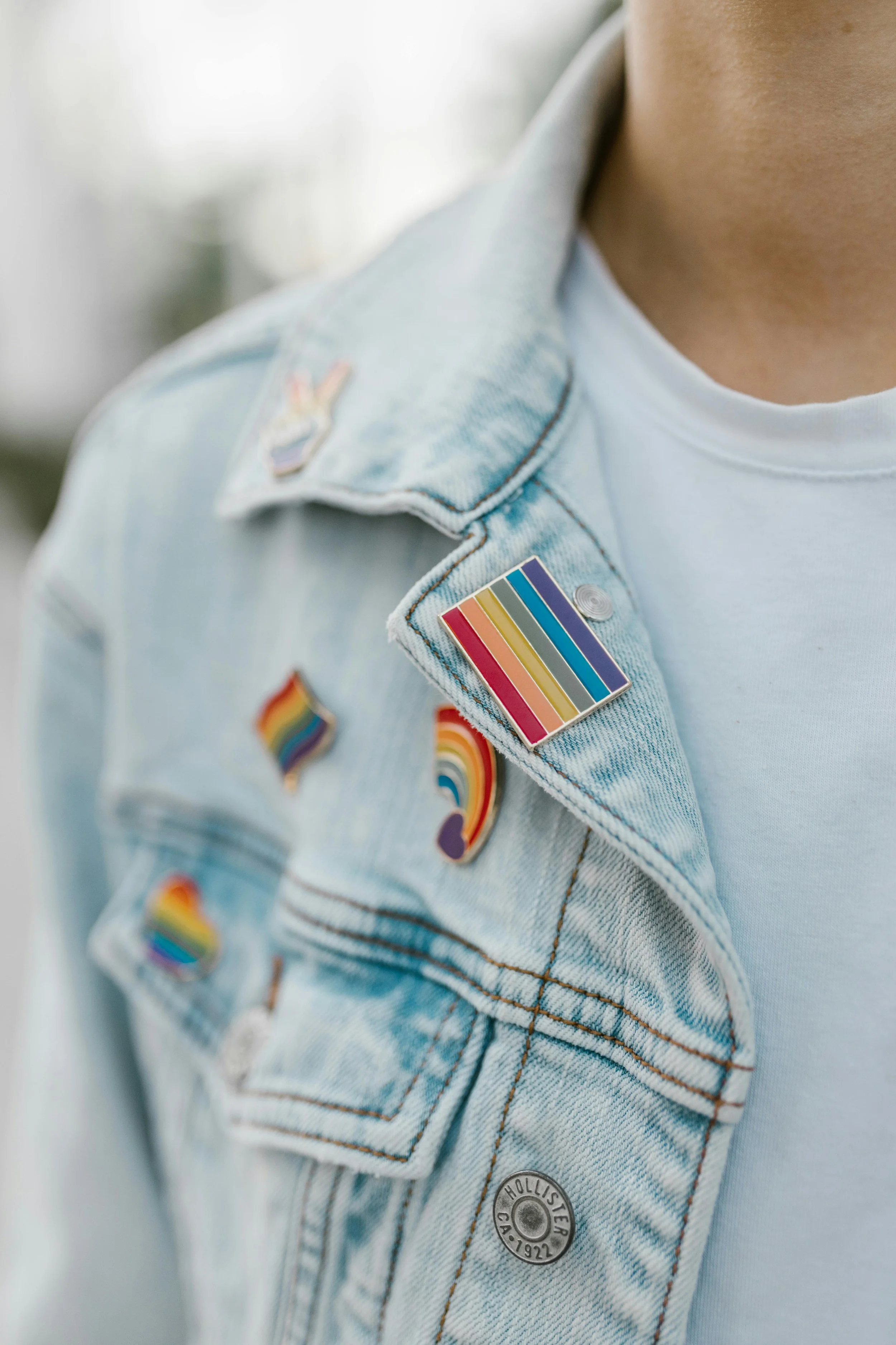 Close-up of a person wearing a light blue denim jacket with rainbow pride pins and a white T-shirt underneath.