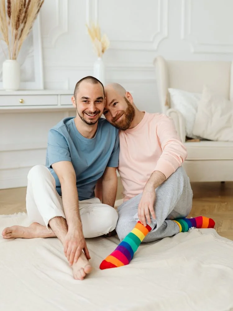 Two male presenting people sitting on the floor leaning into each other and smiling