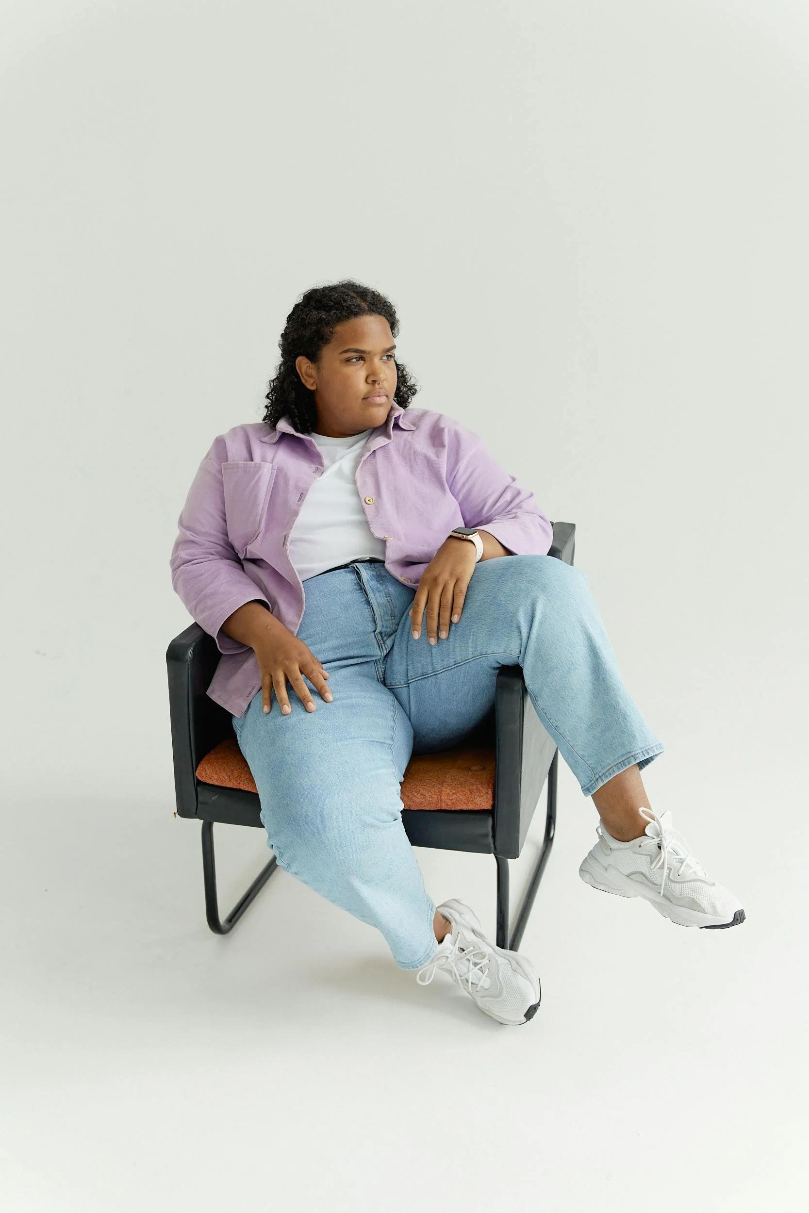 A person sitting in a black chair with orange upholstery, wearing a lavender jacket, white T-shirt, light blue jeans, and white sneakers, against a plain white background.