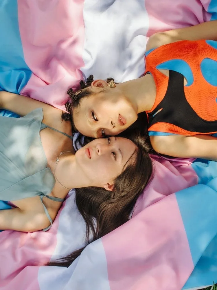 Two people laying on top of a trans pride flag looking at each other.