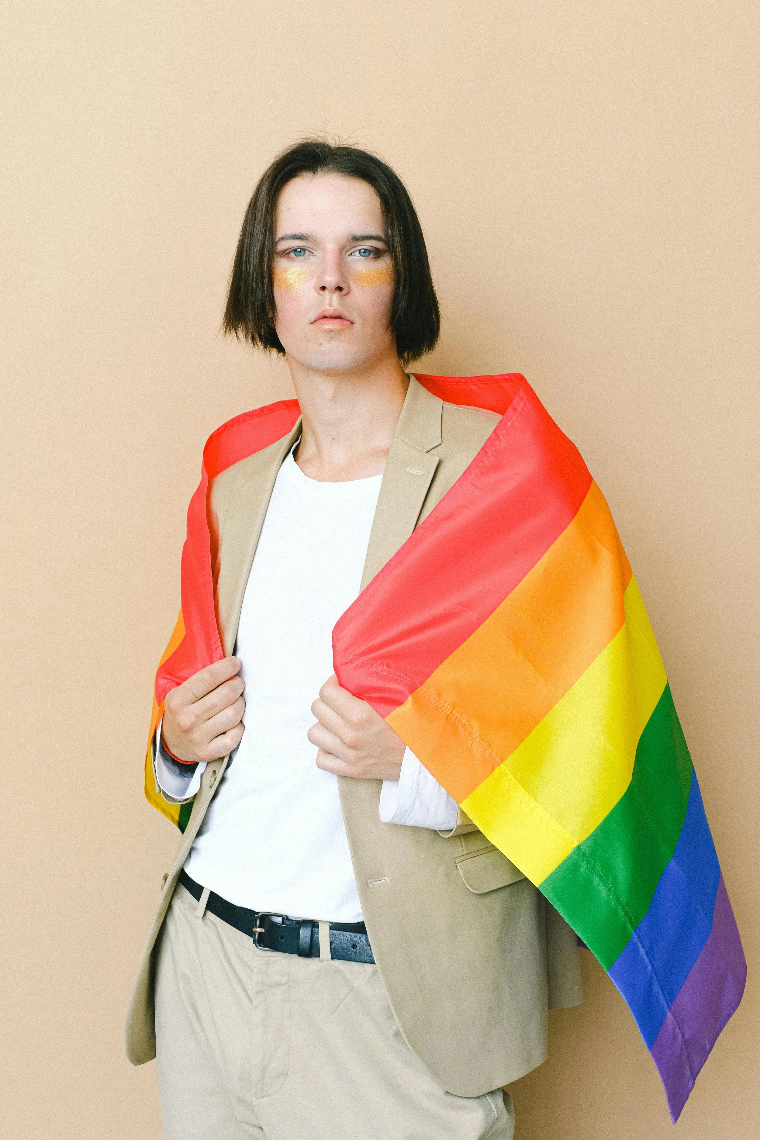 A person with shoulder-length black hair and blue eyes holds a rainbow pride flag over their shoulders, wearing a beige blazer, white shirt, and beige pants against a neutral background.