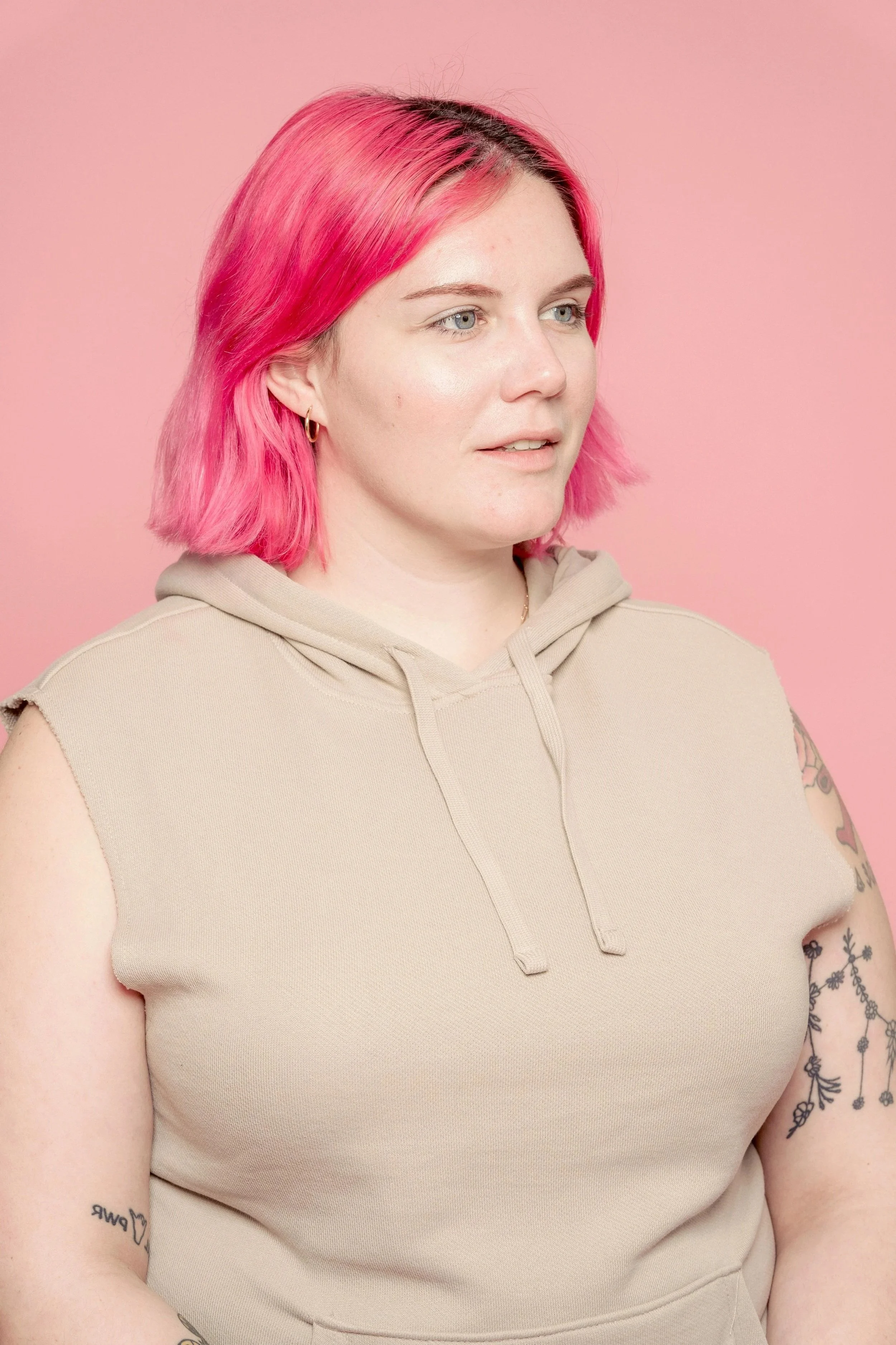 A woman with pink hair wearing a beige sleeveless hoodie, standing against a pink background.
