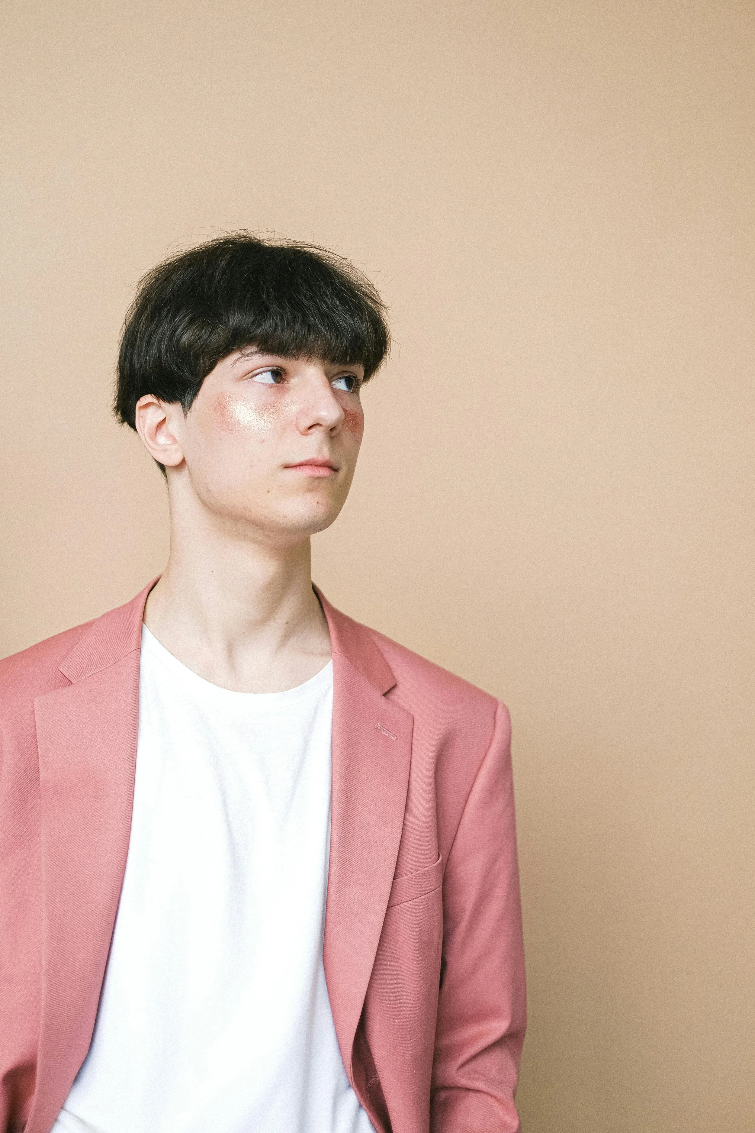 A young person with dark hair and fair skin wearing a pink blazer and white t-shirt, standing against a beige background, looking to the side.
