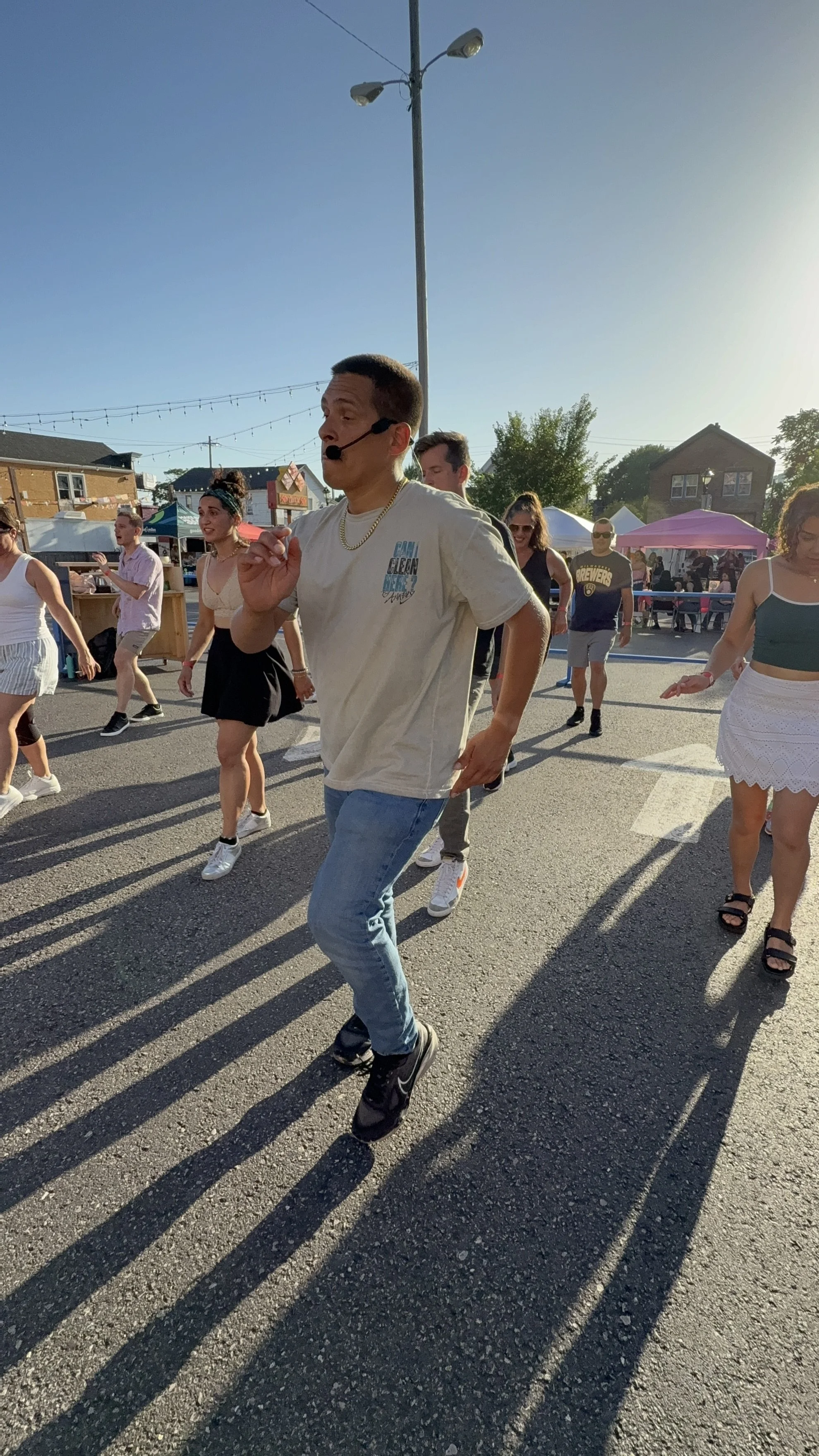 Co-Founder Joshua Burgos wearing a headset microphone and a gold chain, dancing in front of a crowd at an outdoor event during sunset. Tents and people are visible in the background.