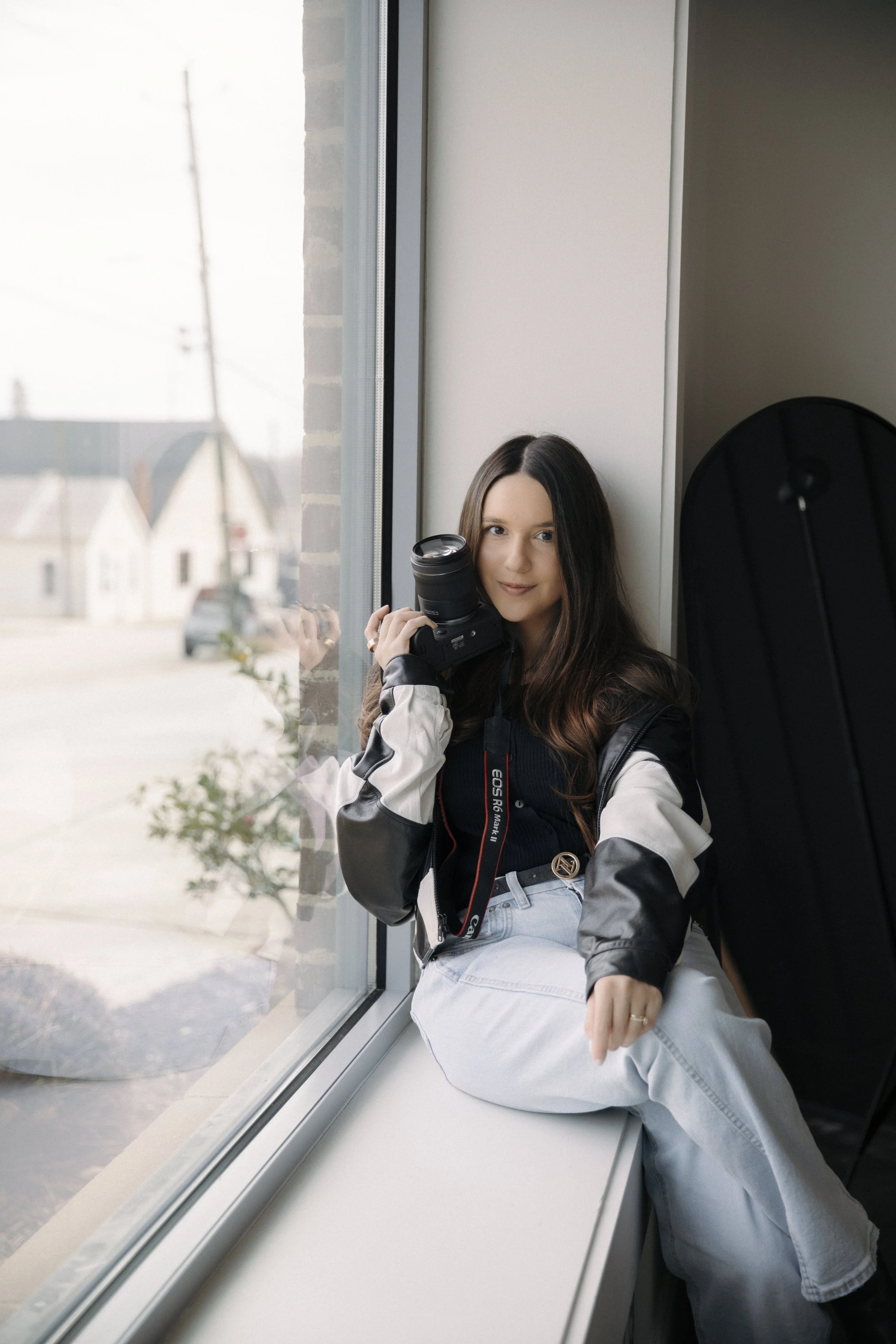 Young woman sitting by a large window holding a camera, with a camera bag nearby.