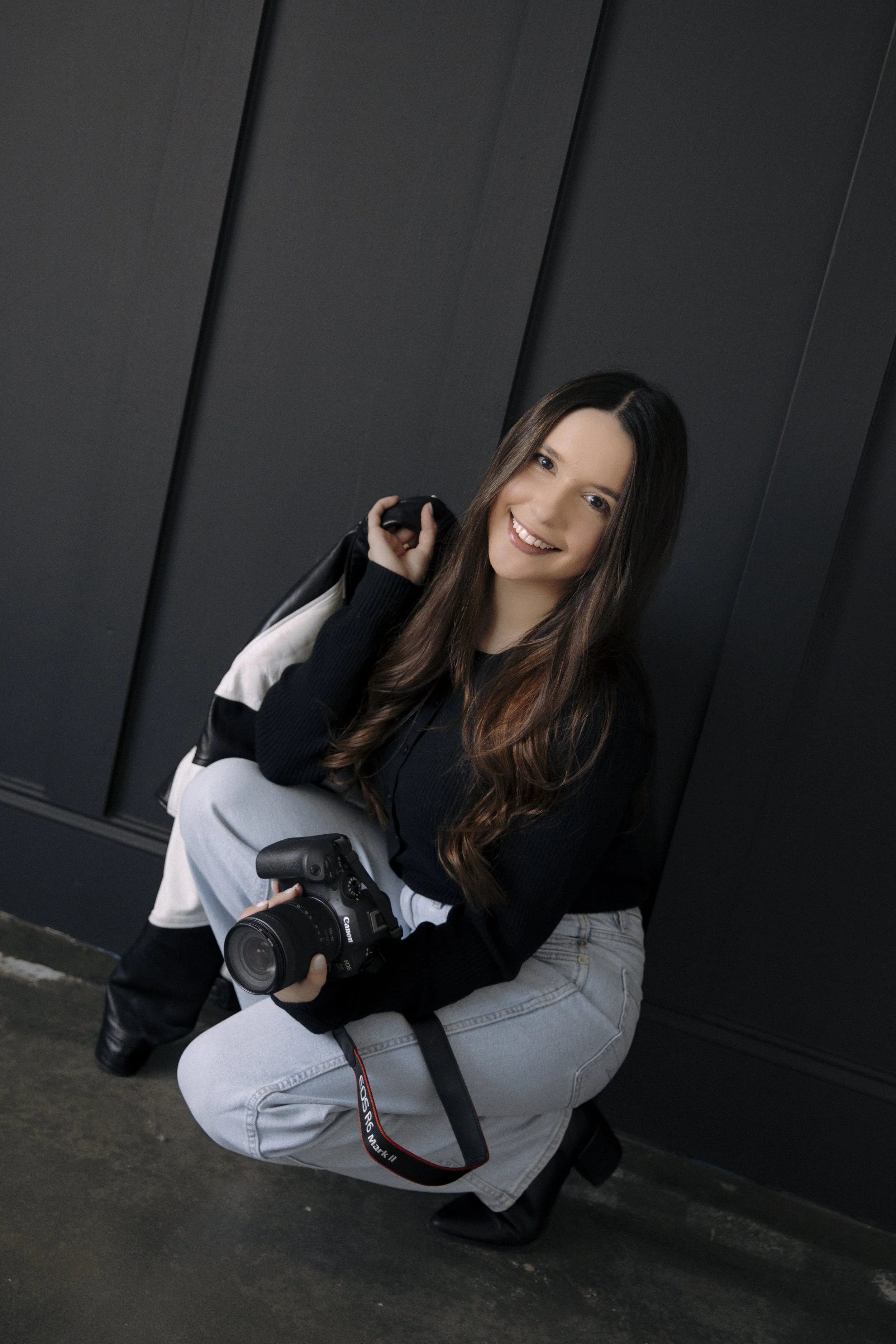 Young woman with long brown hair wearing a black sweater and light-colored jeans, holding a professional camera, smiling and crouching against a dark wall.