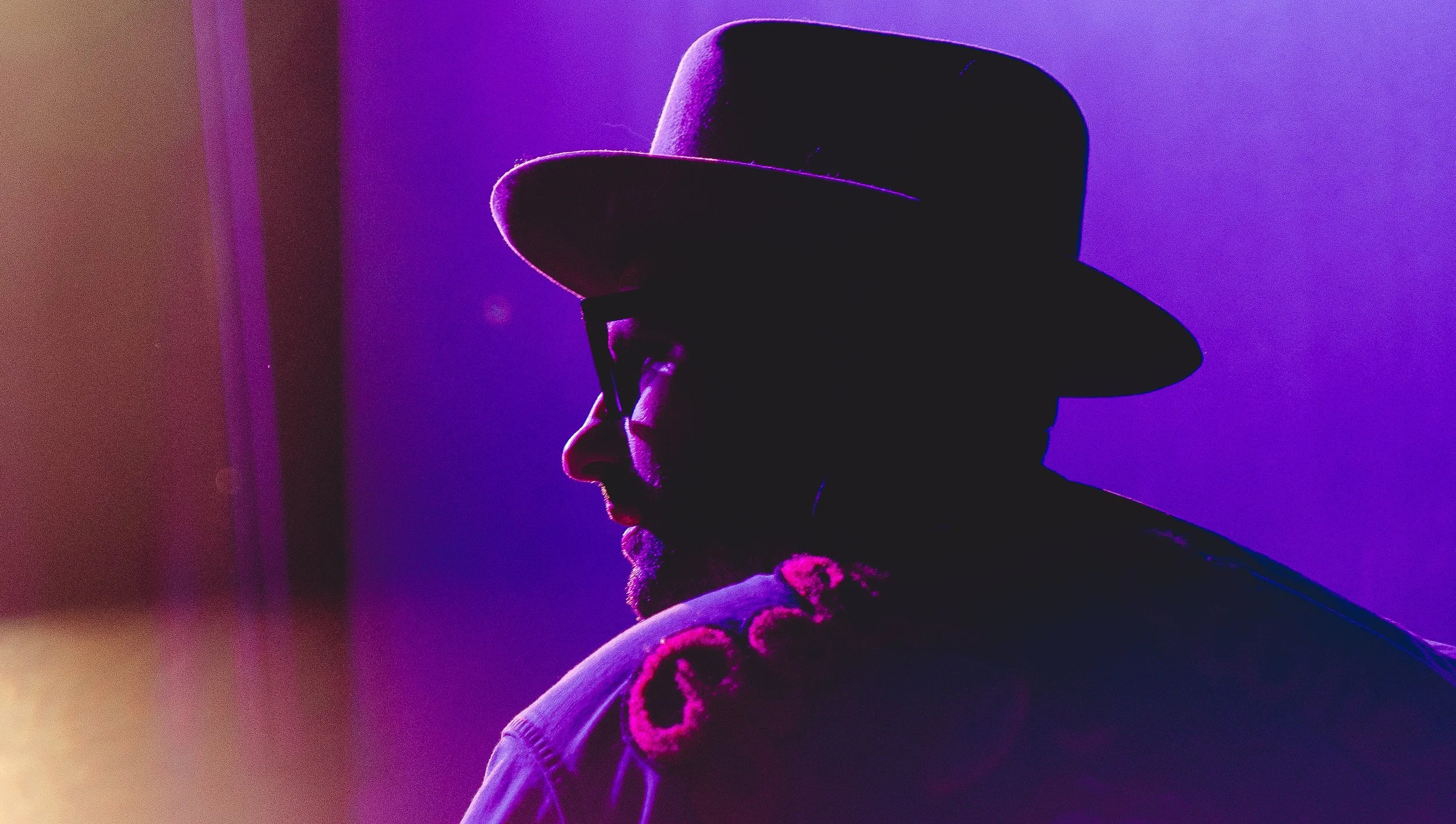 Silhouette of a person wearing a hat and glasses, backlit with purple and pink lighting, looking downward.