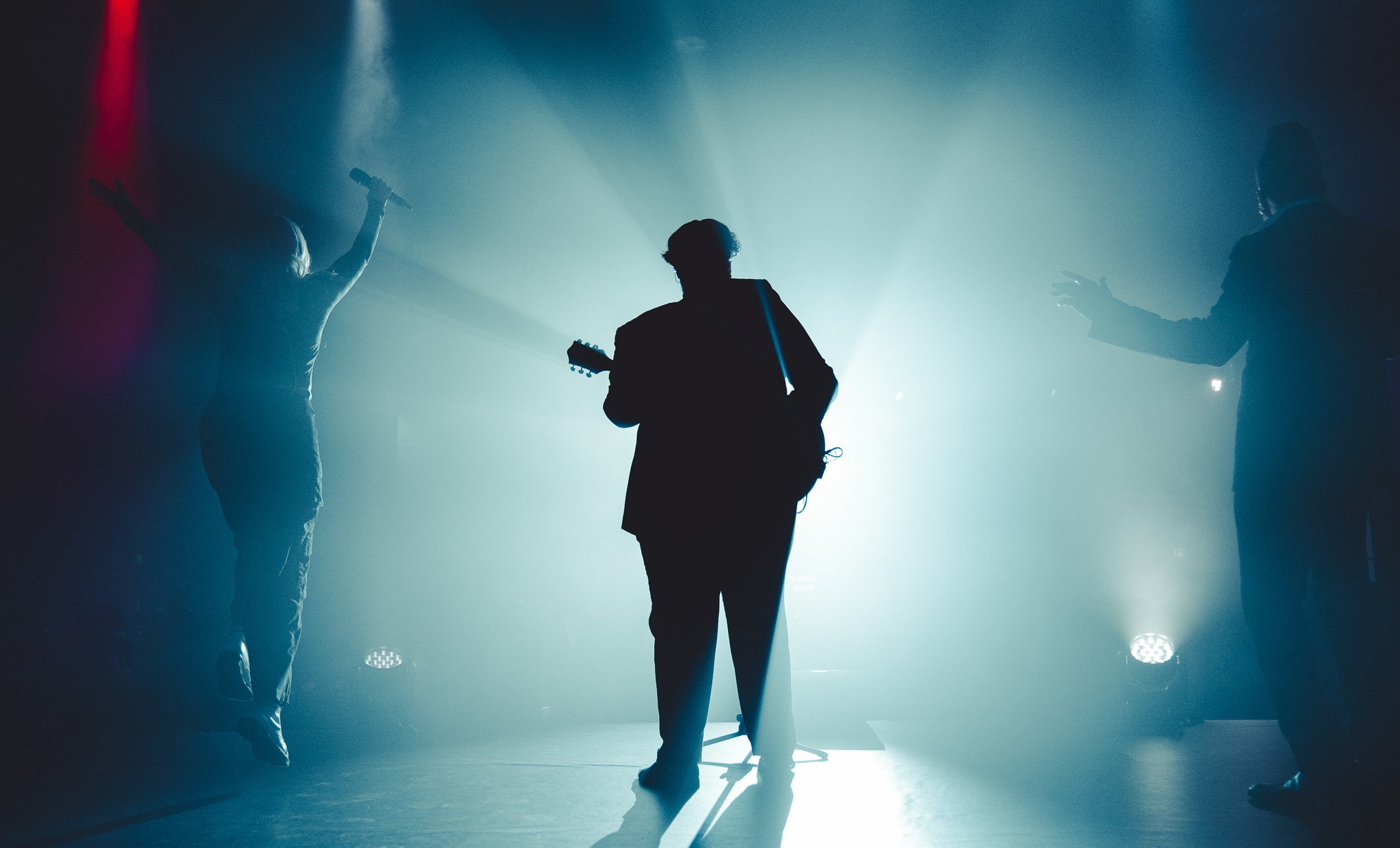 Silhouettes of three people on stage, one playing guitar, others singing or performing, with bright stage lights creating a dramatic atmosphere.