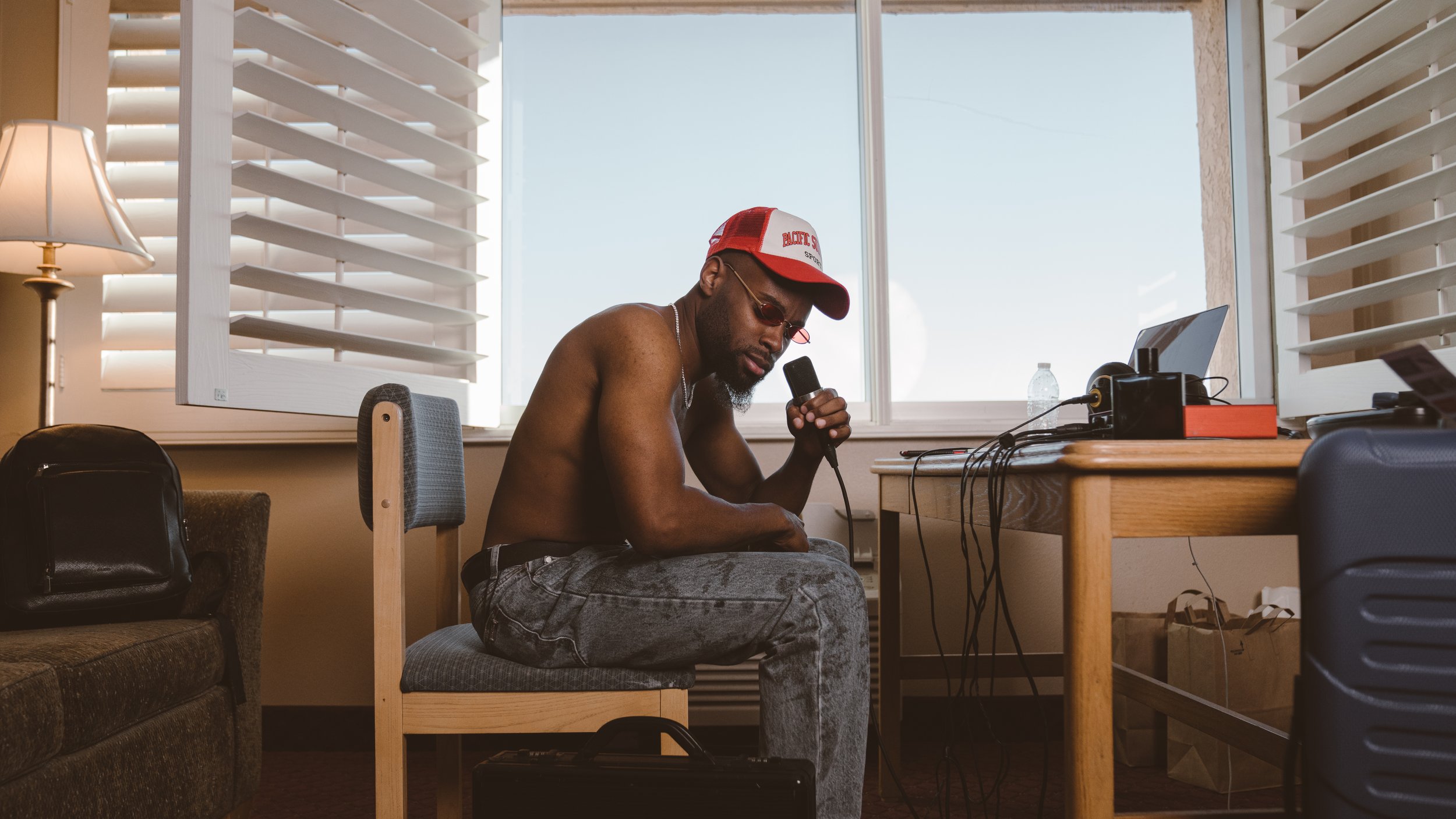 A shirtless man wearing sunglasses, a red and white baseball cap, and gray jeans, sitting on a chair in a room with bright sunlight outside, holding a microphone while looking down at electronic equipment on a wooden desk.