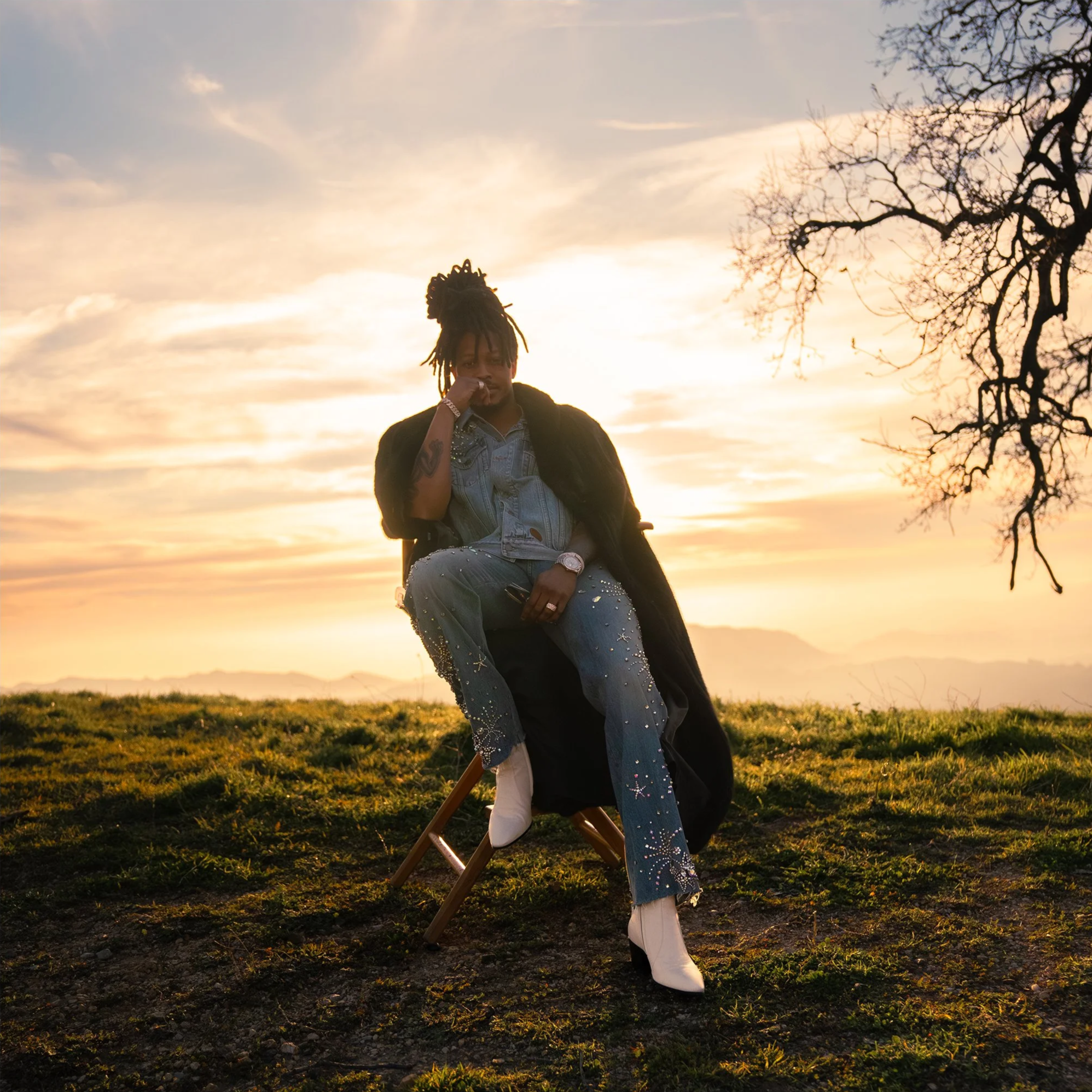Person sitting on a wooden chair outdoors during sunset, wearing jeans with embellishments, white boots, denim shirt, and black coat, with a tree and distant mountains in the background.