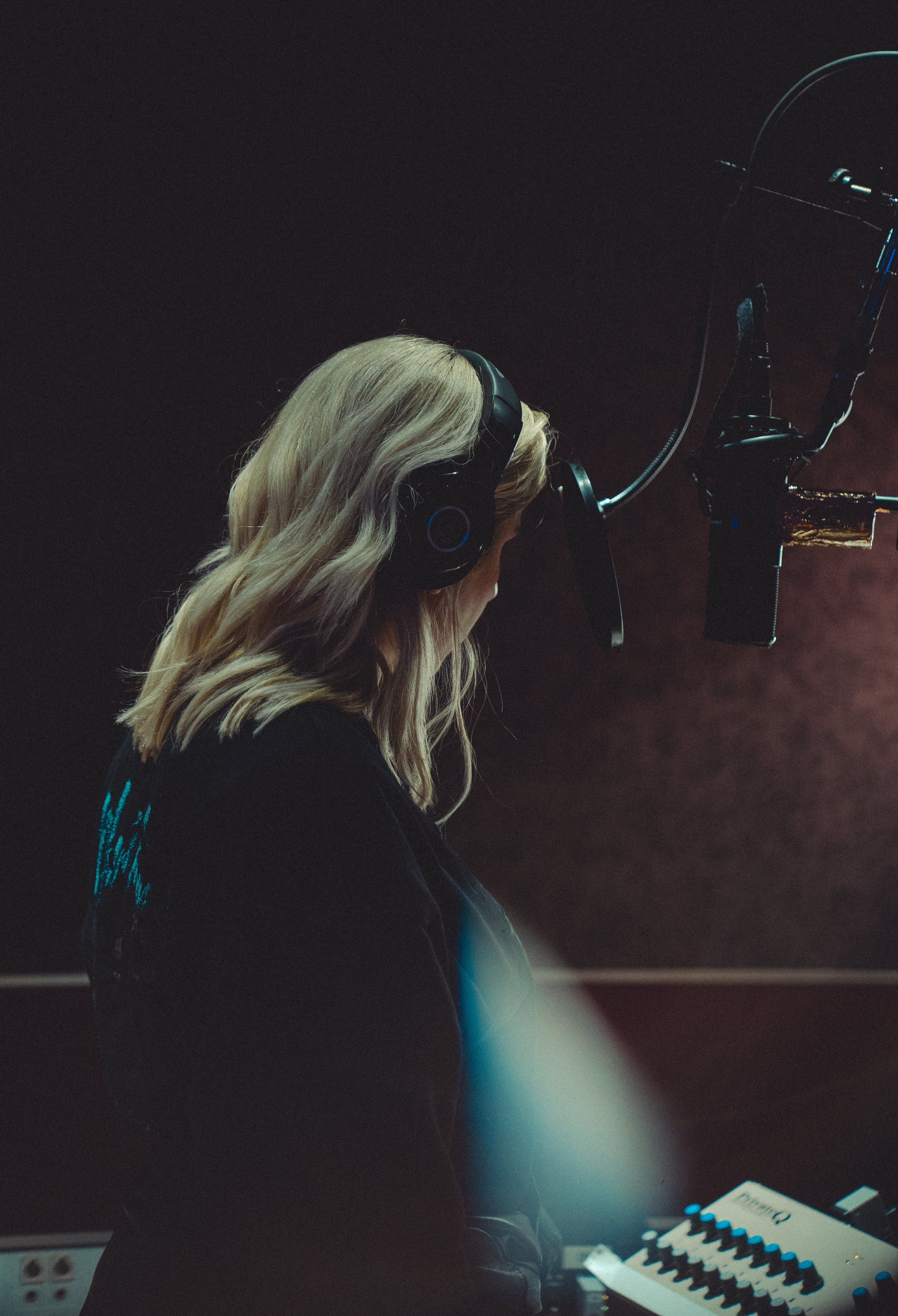 A woman with blonde hair wearing headphones is singing or speaking into a microphone in a recording studio.