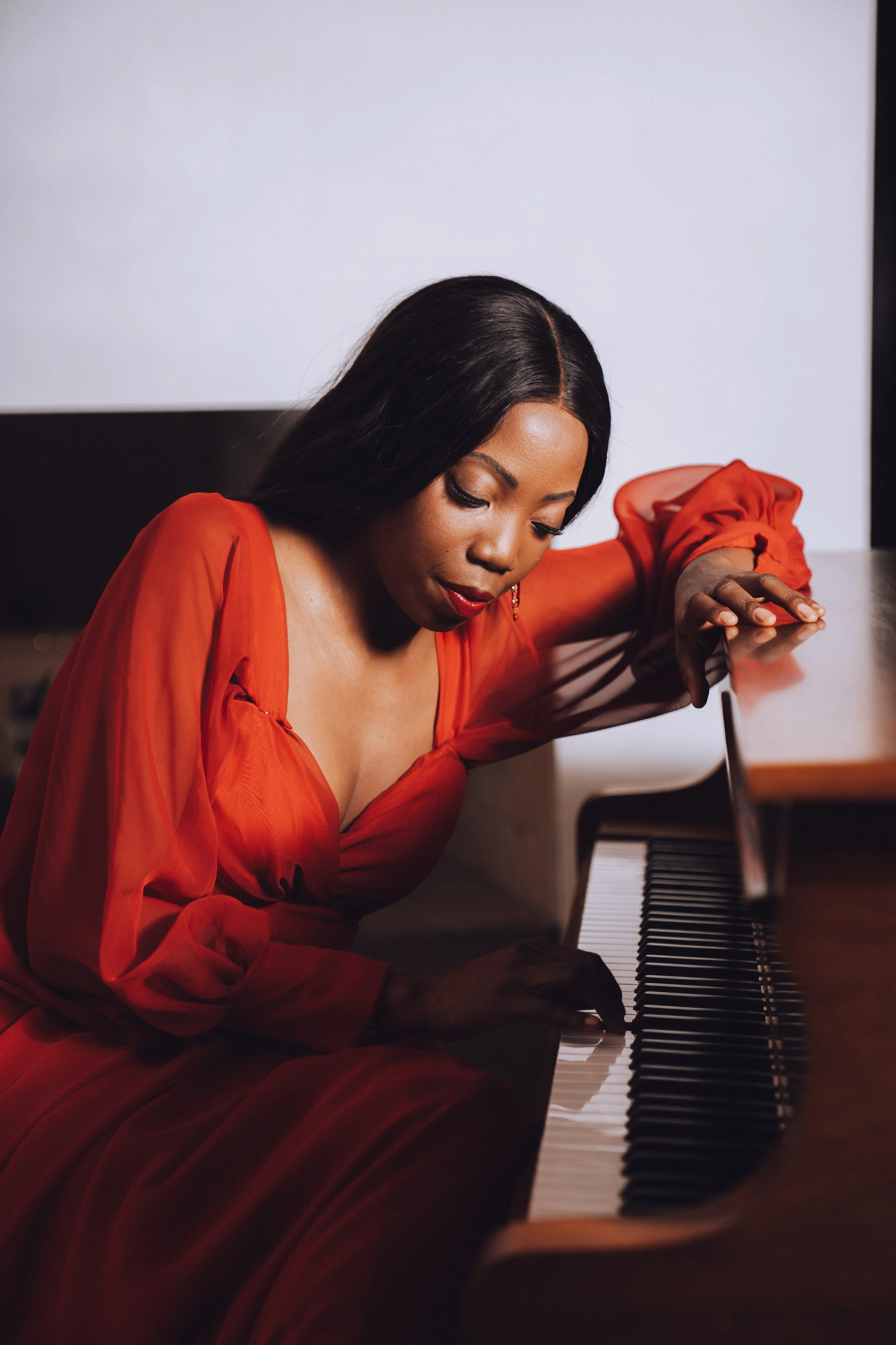 A woman in a red dress leaning over a grand piano, with her hand on the keys and her head bowed, appearing to be focused or expressing emotion while playing.
