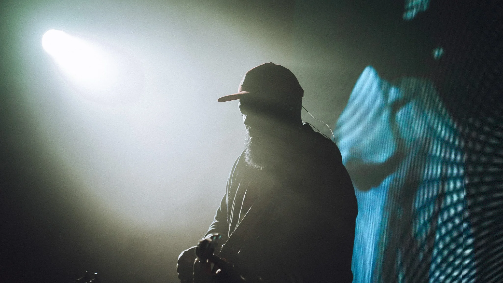 Silhouette of a DJ wearing a baseball cap, with a large screen displaying abstract visuals in the background, and stage lighting creating a dramatic effect.