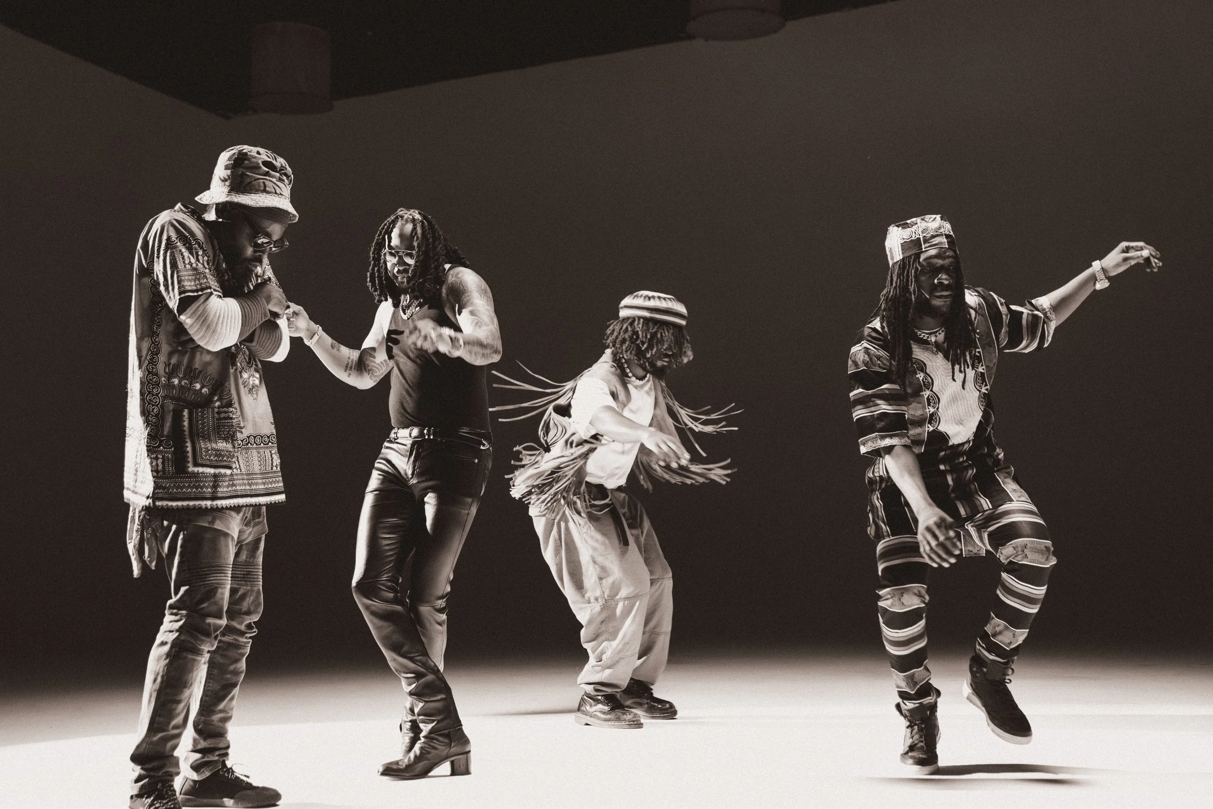 Four people dancing on stage in black and white, dressed in casual and traditional clothing, with energetic poses and movement.