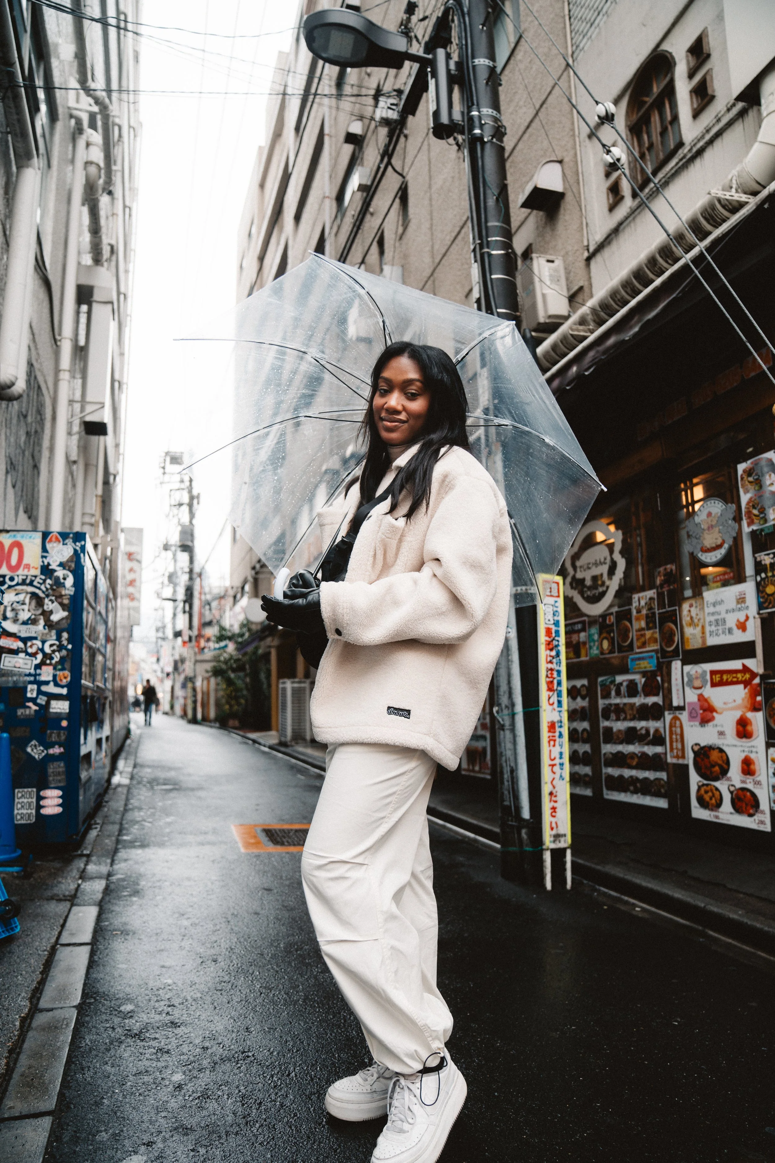 A woman holding a transparent umbrella standing in a wet city alleyway, wearing a cream fleece jacket, beige pants, and white sneakers, with buildings, a restaurant, and power lines in the background.