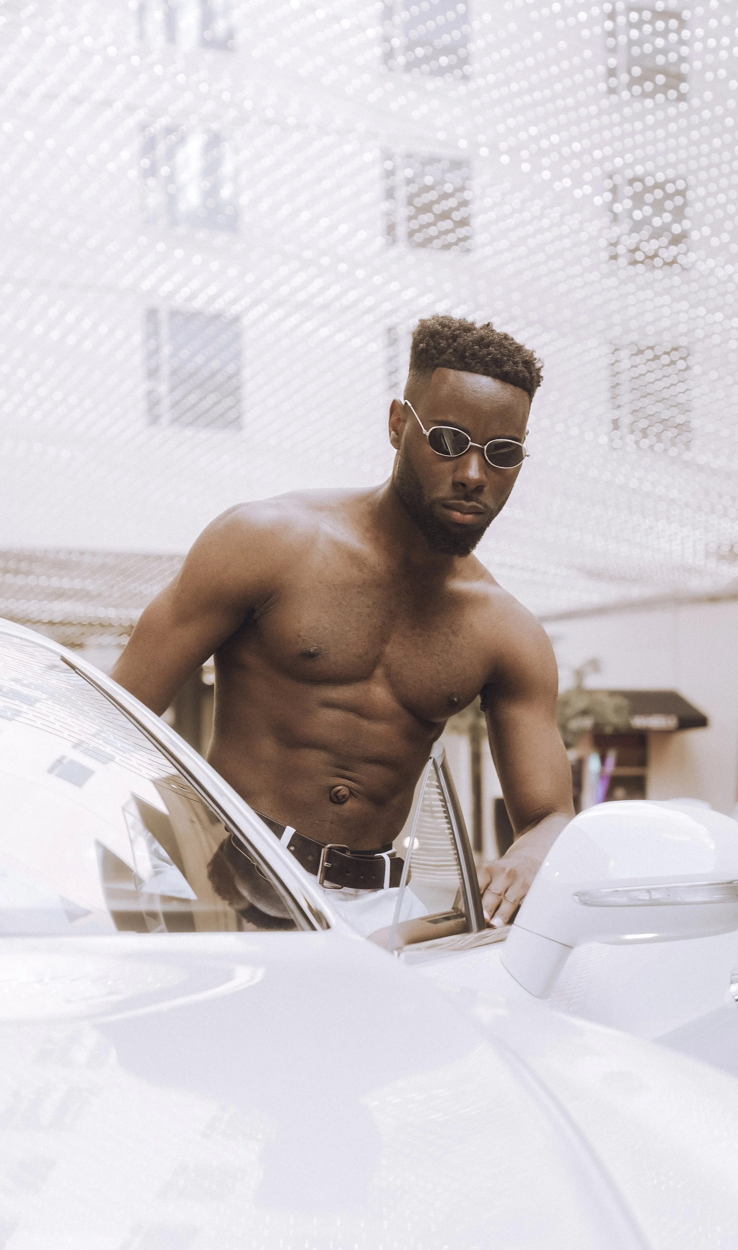 A shirtless man with glasses leaning over a white car in a modern, artistic environment.