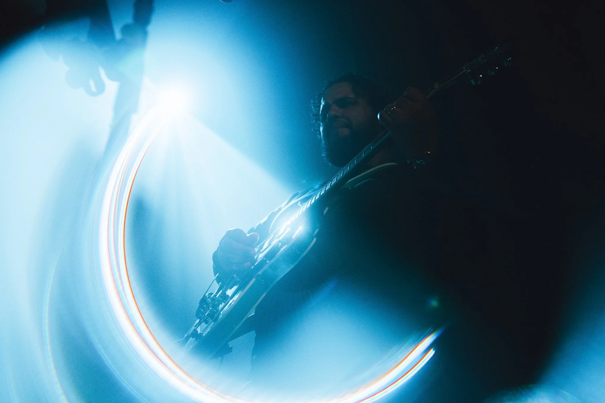 A musician plays an electric guitar on stage illuminated by blue stage lights with lens flare effects.