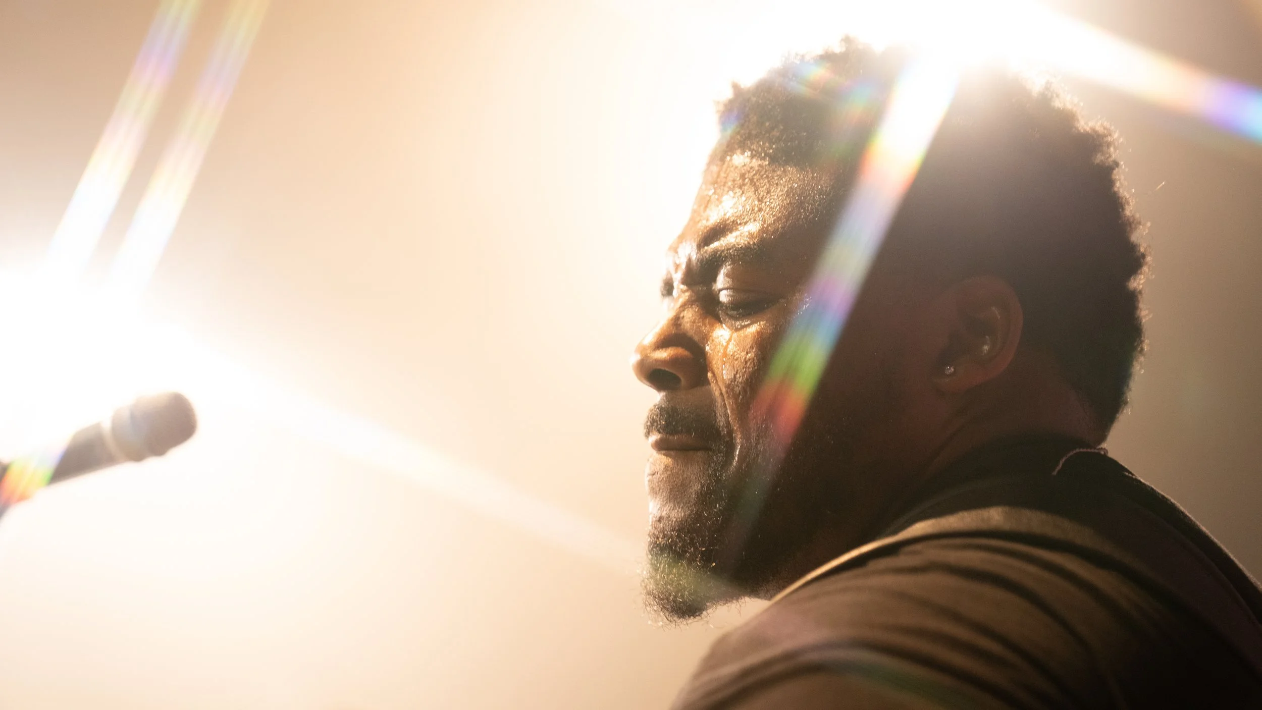 A man with short curly hair and earrings, eyes closed, facing the bright light source with rainbow lens flare effects.