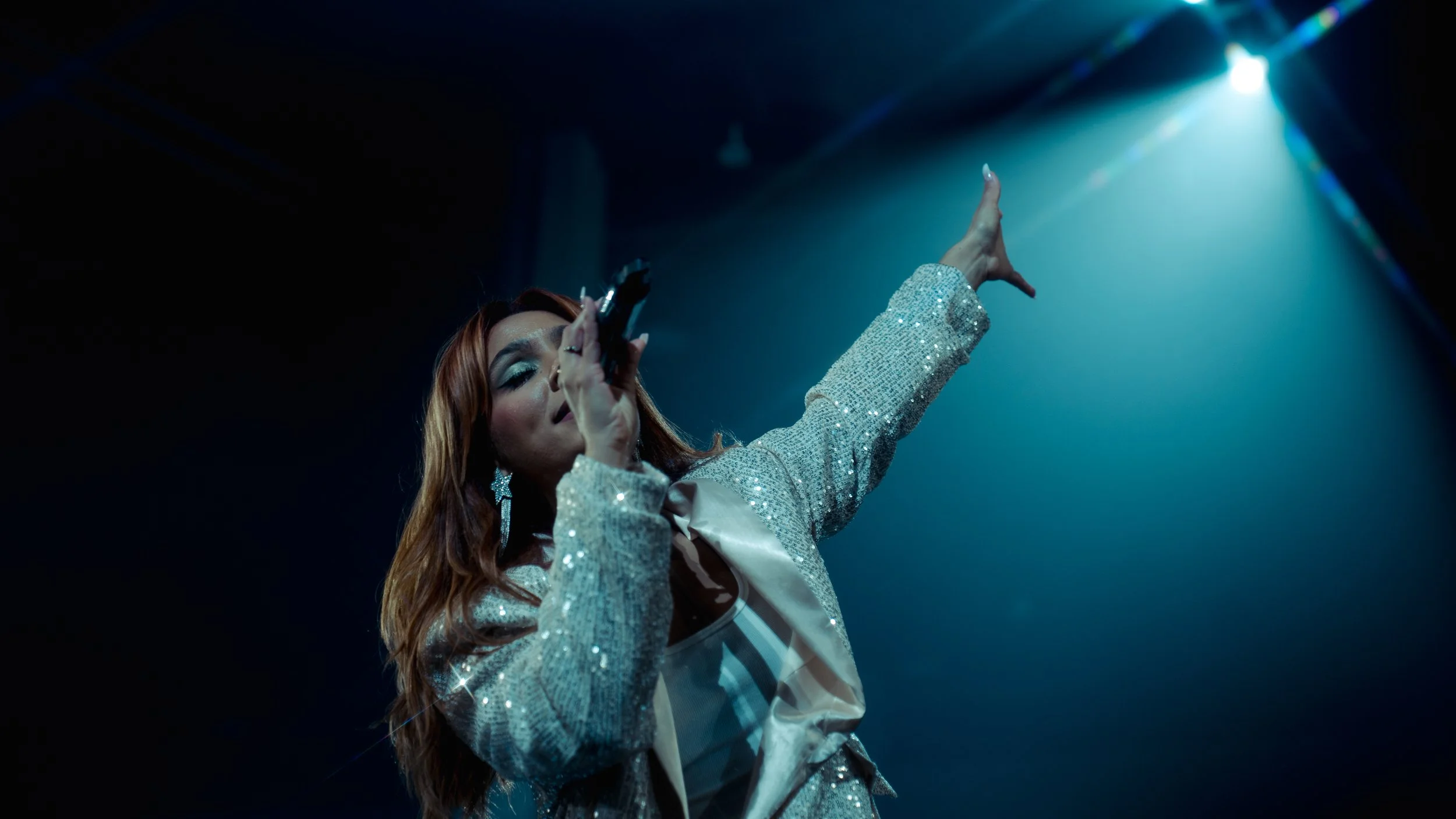 Performer singing into a microphone on stage, wearing a sparkly silver jacket, with blue stage lighting and a dark background.