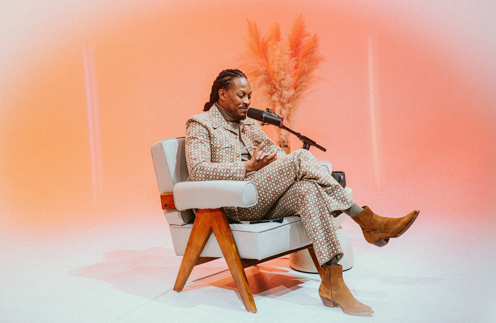 A man with dreadlocks wearing a patterned suit and brown boots sitting in a modern armchair, speaking into a microphone on a stand, with a pinkish background and dried plants in the background.