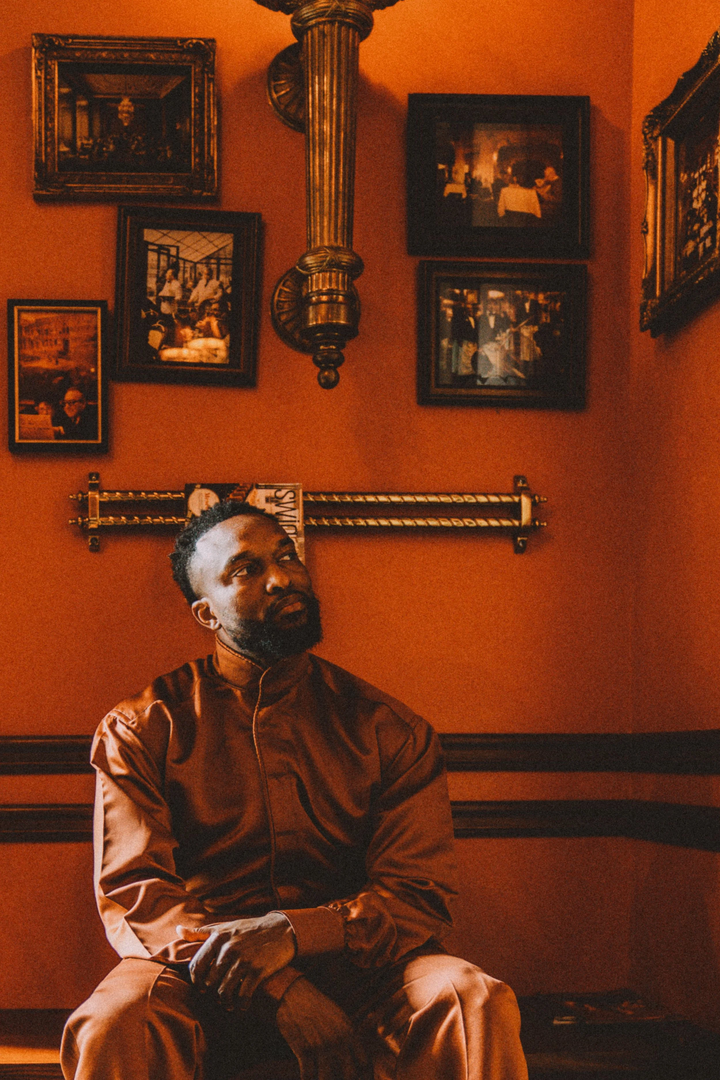 A man sitting on a wooden bench in a room with dark orange walls. The man has a beard, short hair, and is wearing a shiny brown jacket and matching pants. The room has framed photographs on the wall, a large ornate mirror, and a decorative brass wall
