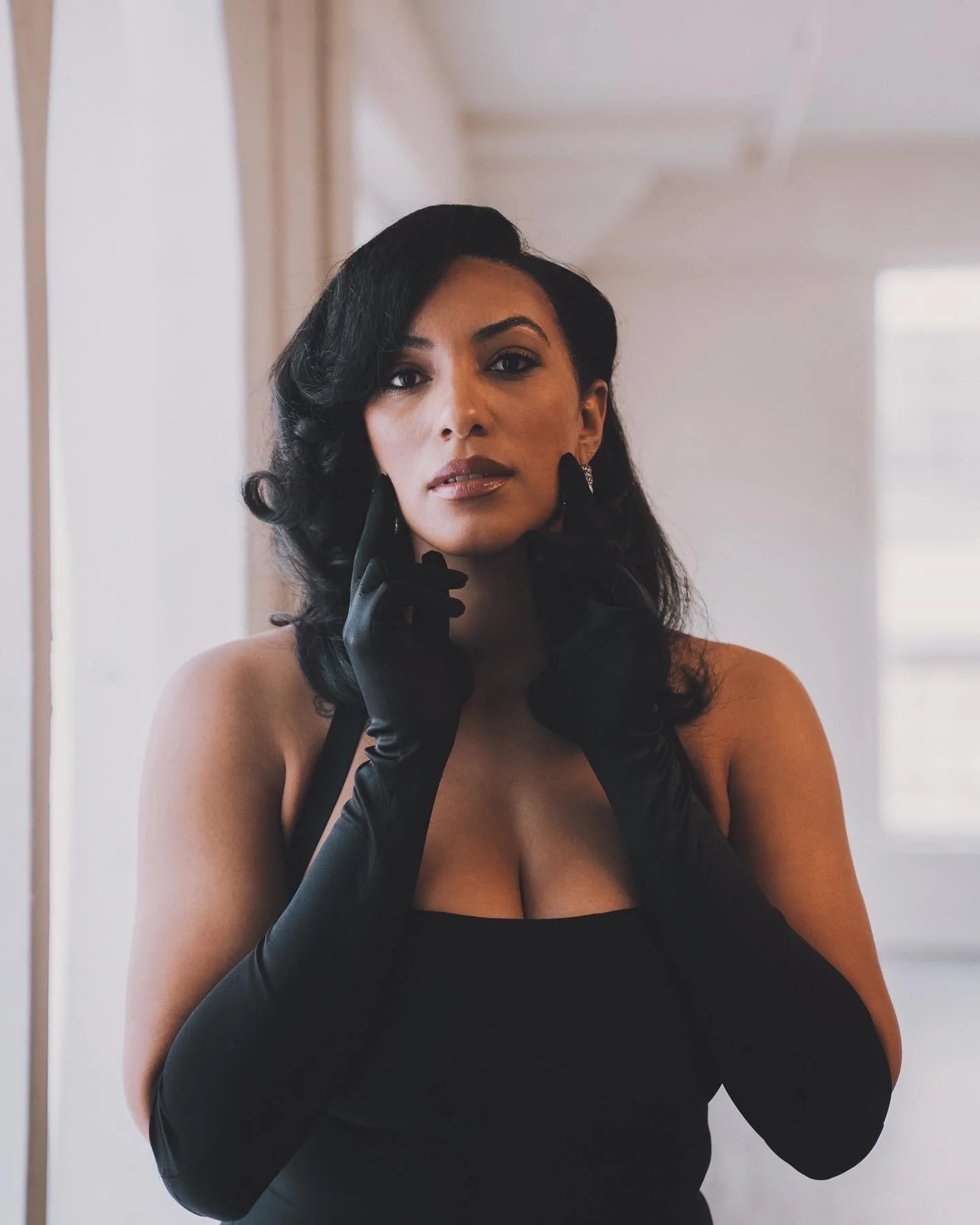 A woman with black, styled hair wearing a black dress and long black gloves, standing indoors with a neutral expression.