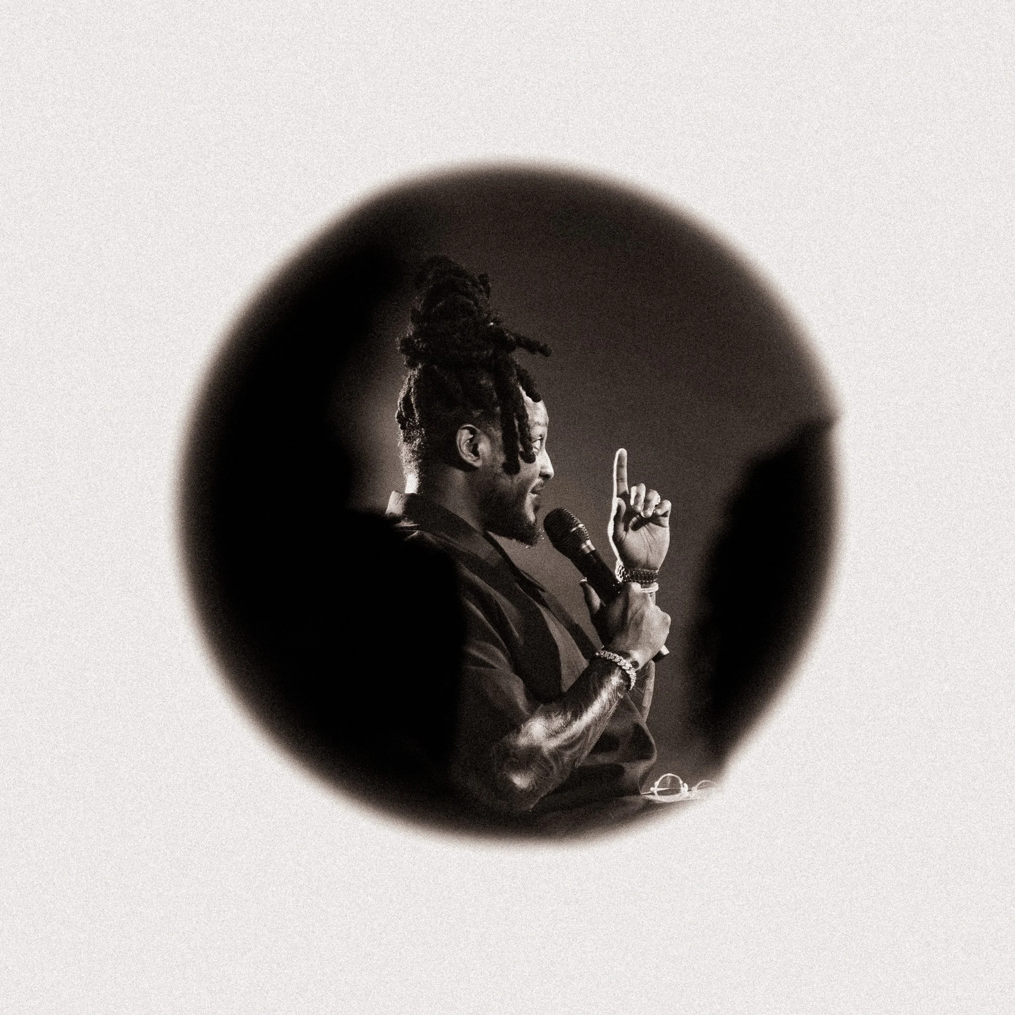 A black and white photo of a man with dreadlocks speaking into a microphone, gesturing with his hand, seen through a circular cutout.