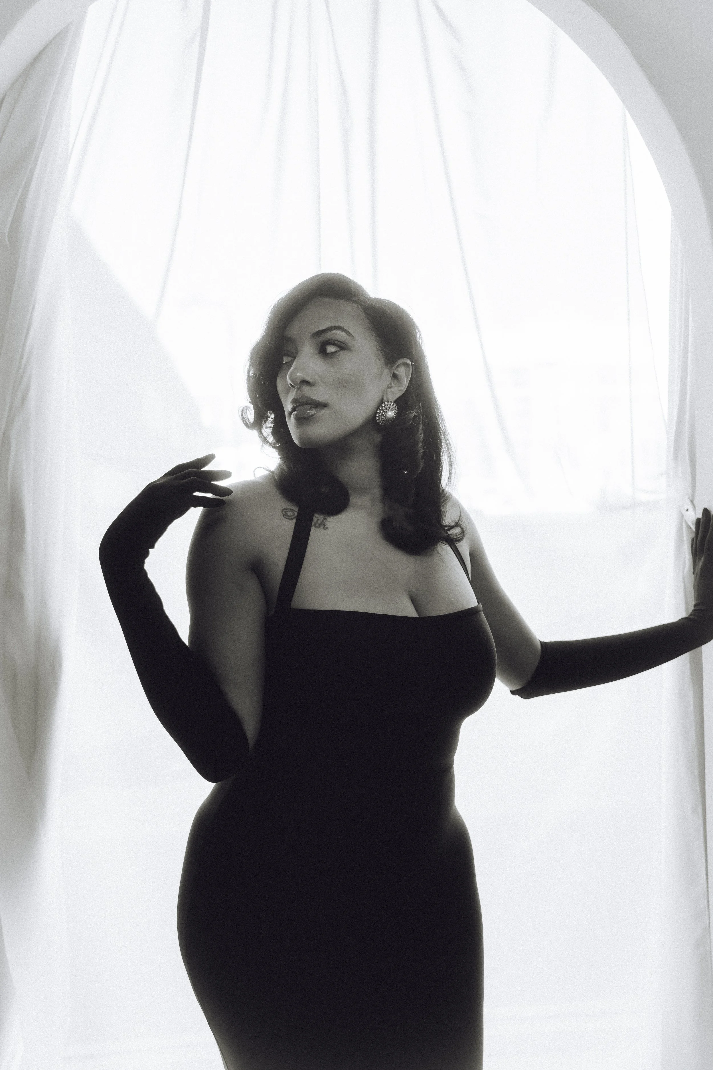A woman in a black dress and long gloves posings beside sheer curtains with backlighting.