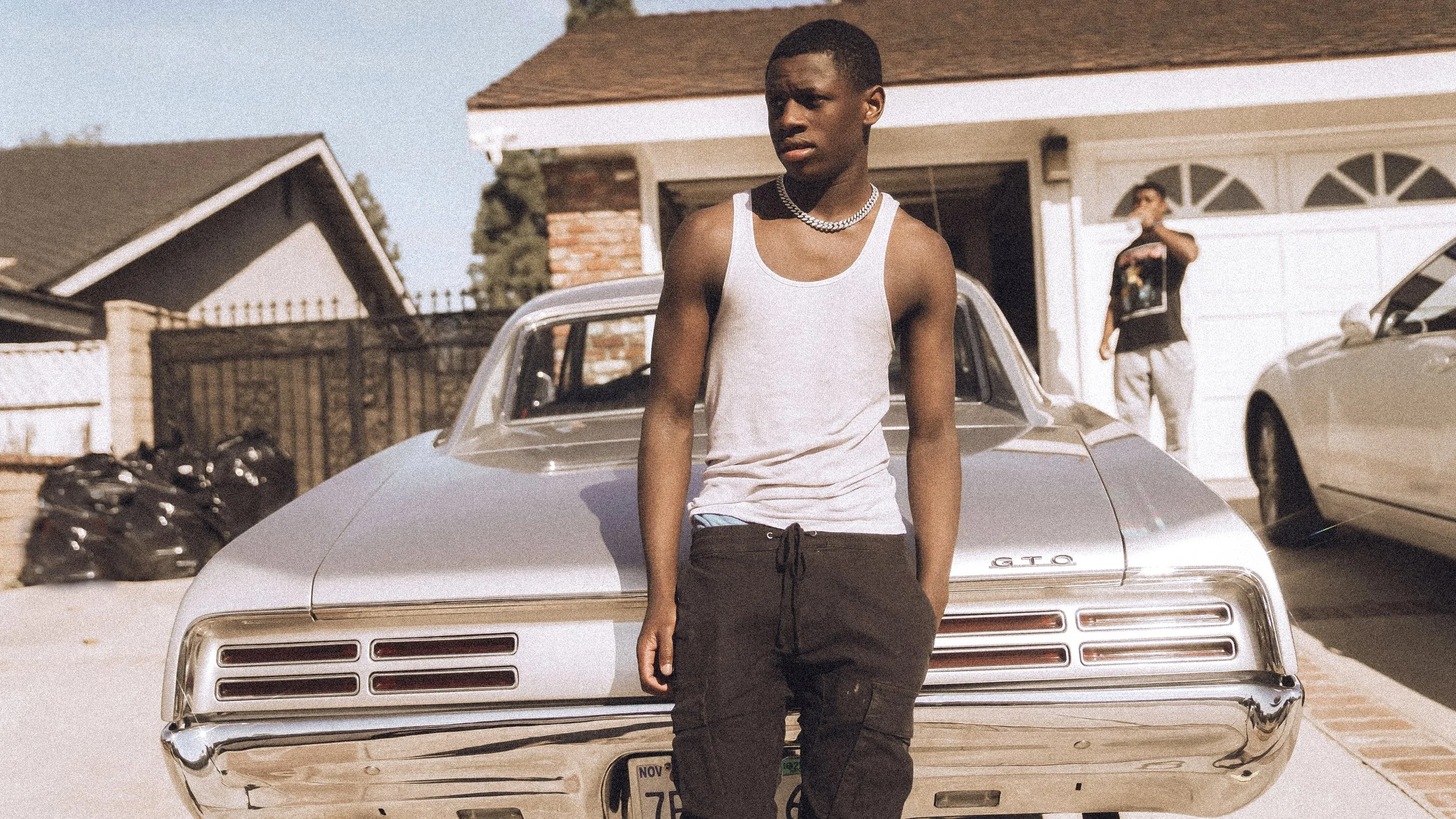 A young man with dark skin and short hair leans against a silver vintage Pontiac GTO car in a residential driveway during daytime. He wears a gray tank top, black drawstring pants, and a chain necklace. There is another person in the background stand