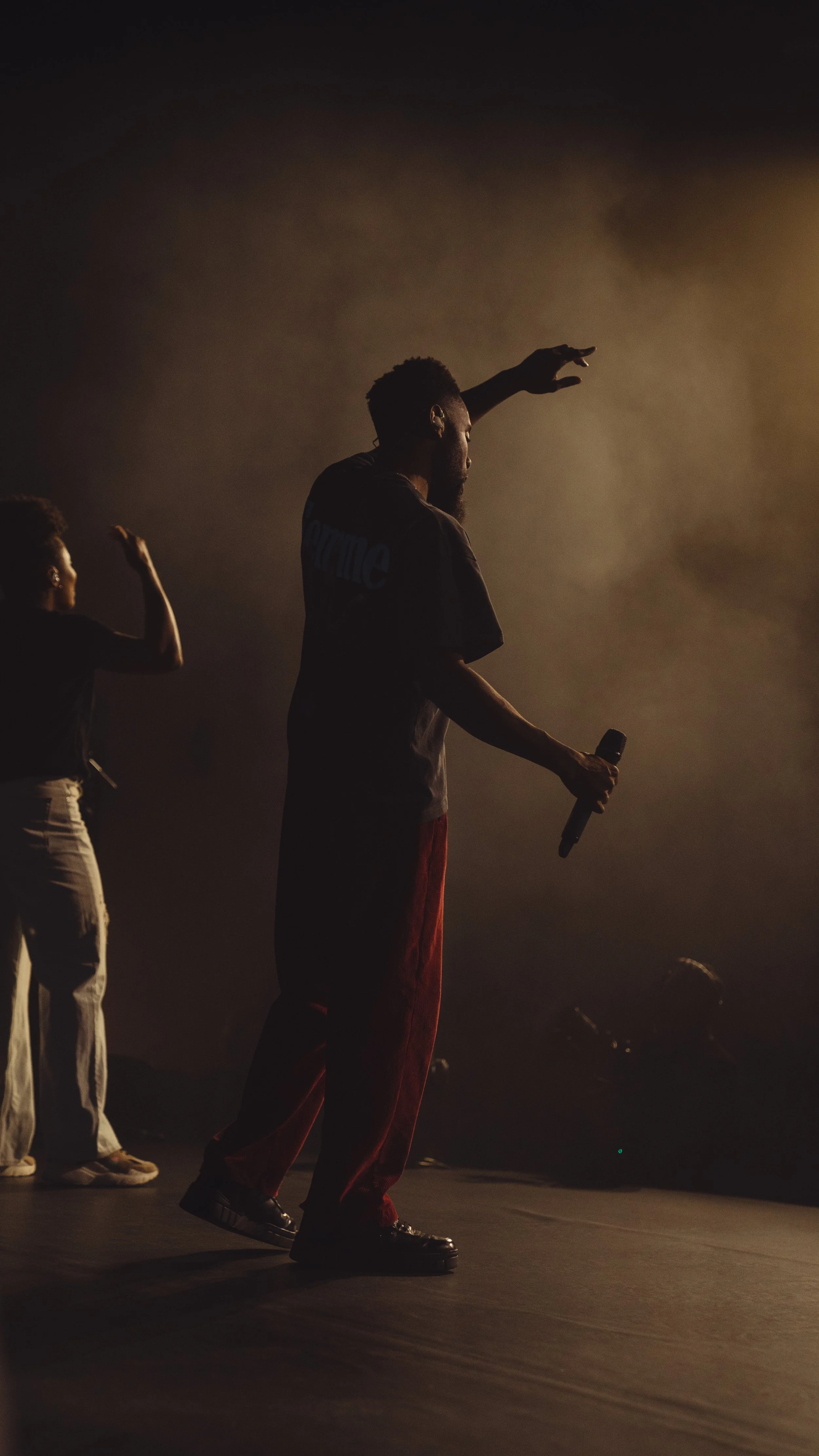 A performer holding a microphone on stage, with another person dancing in the background, illuminated by warm stage lighting.