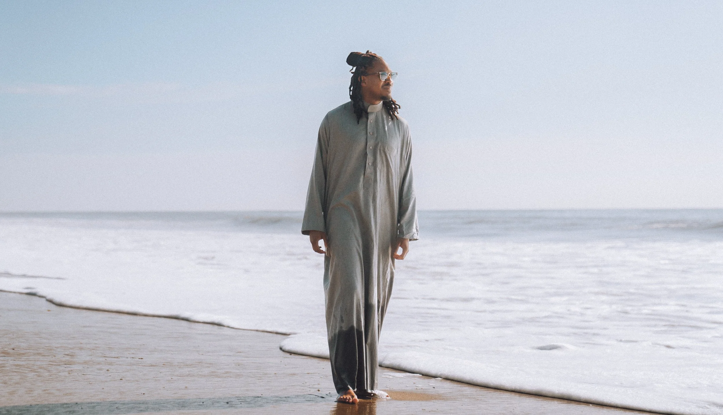 A person with dreadlocks, wearing glasses and a gray onesie, standing on the beach near the water.