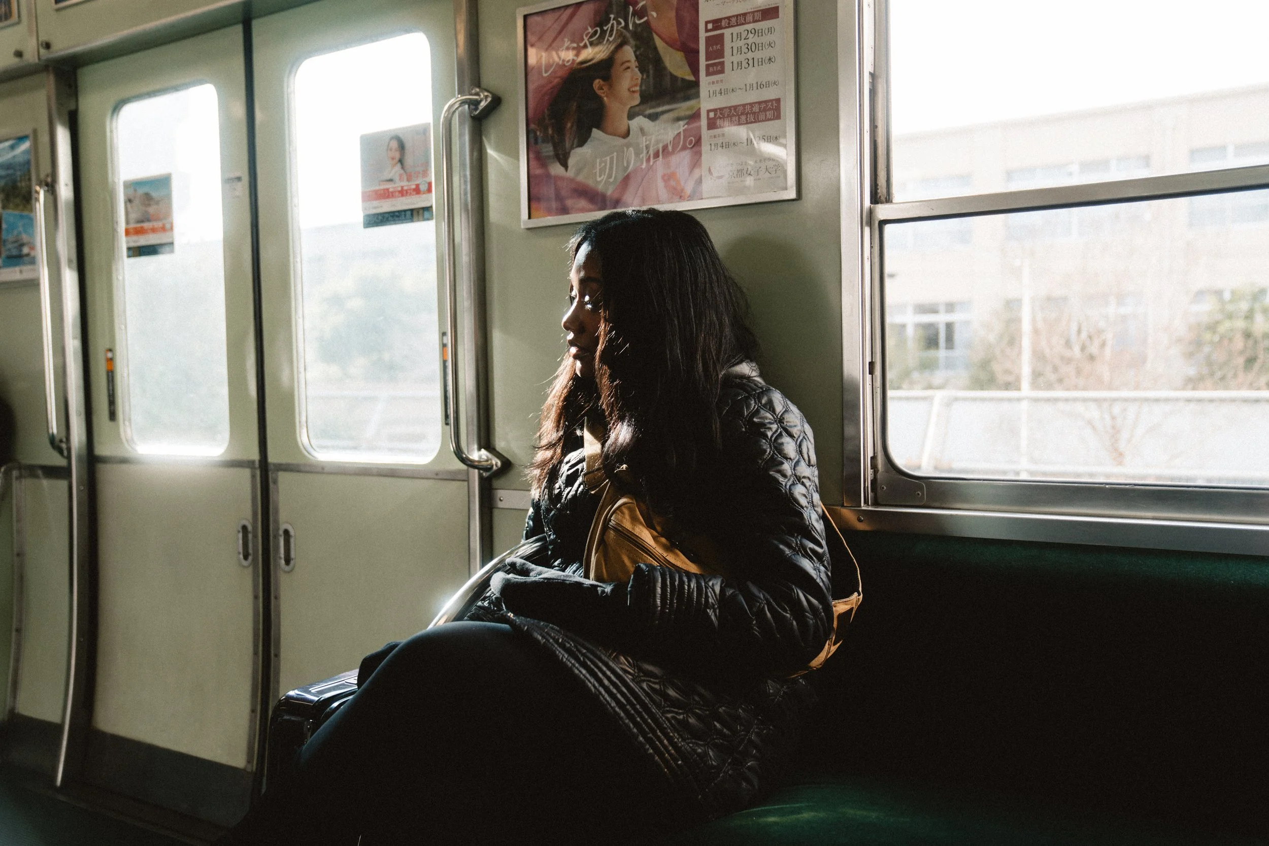 A woman with dark hair wearing a black quilted jacket and holding a yellow purse, sitting on a train with a luggage bag beside her, looking out the window.