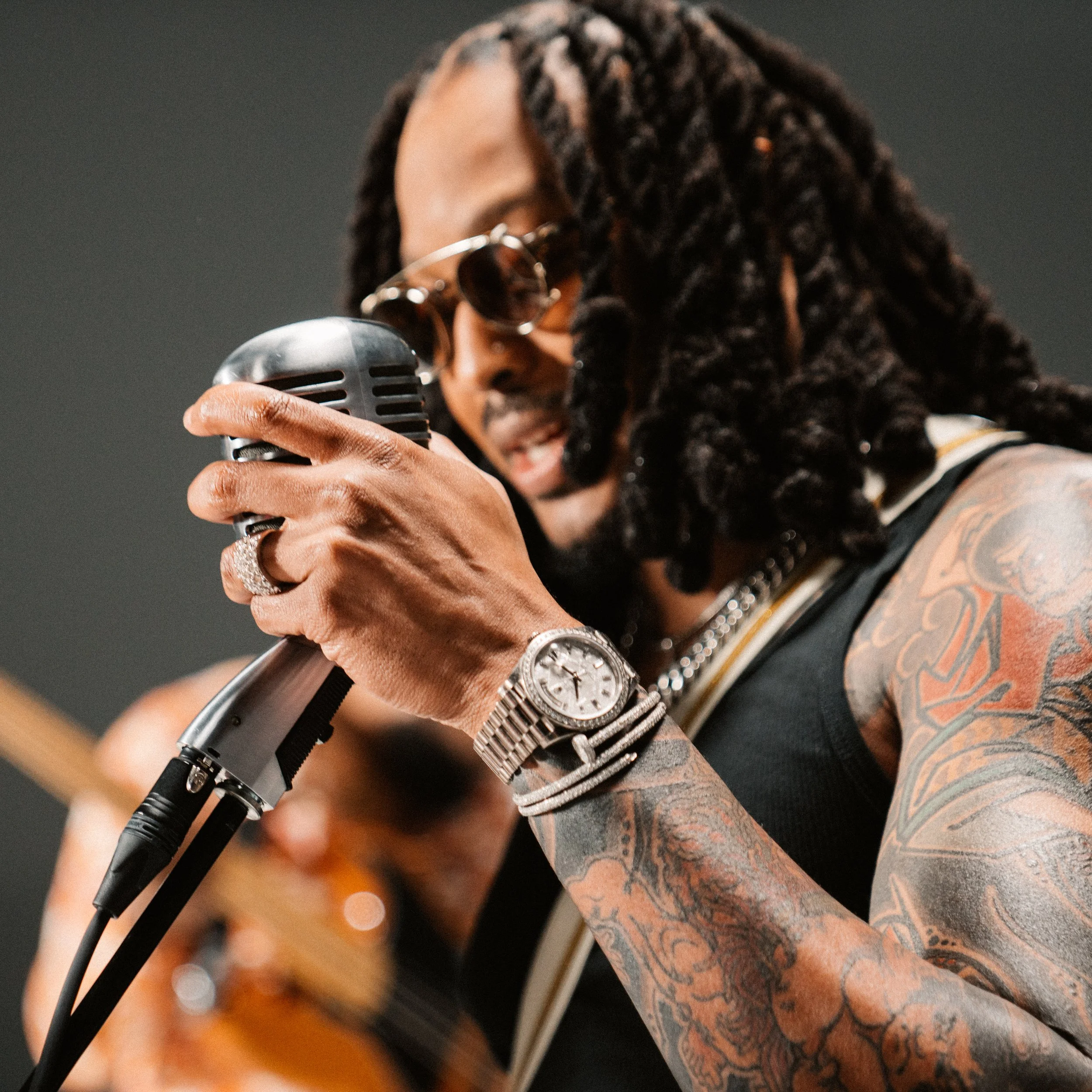 A man with dreadlocks and tattoos singing into a vintage microphone while wearing sunglasses, jewelry, and a sleeveless shirt.