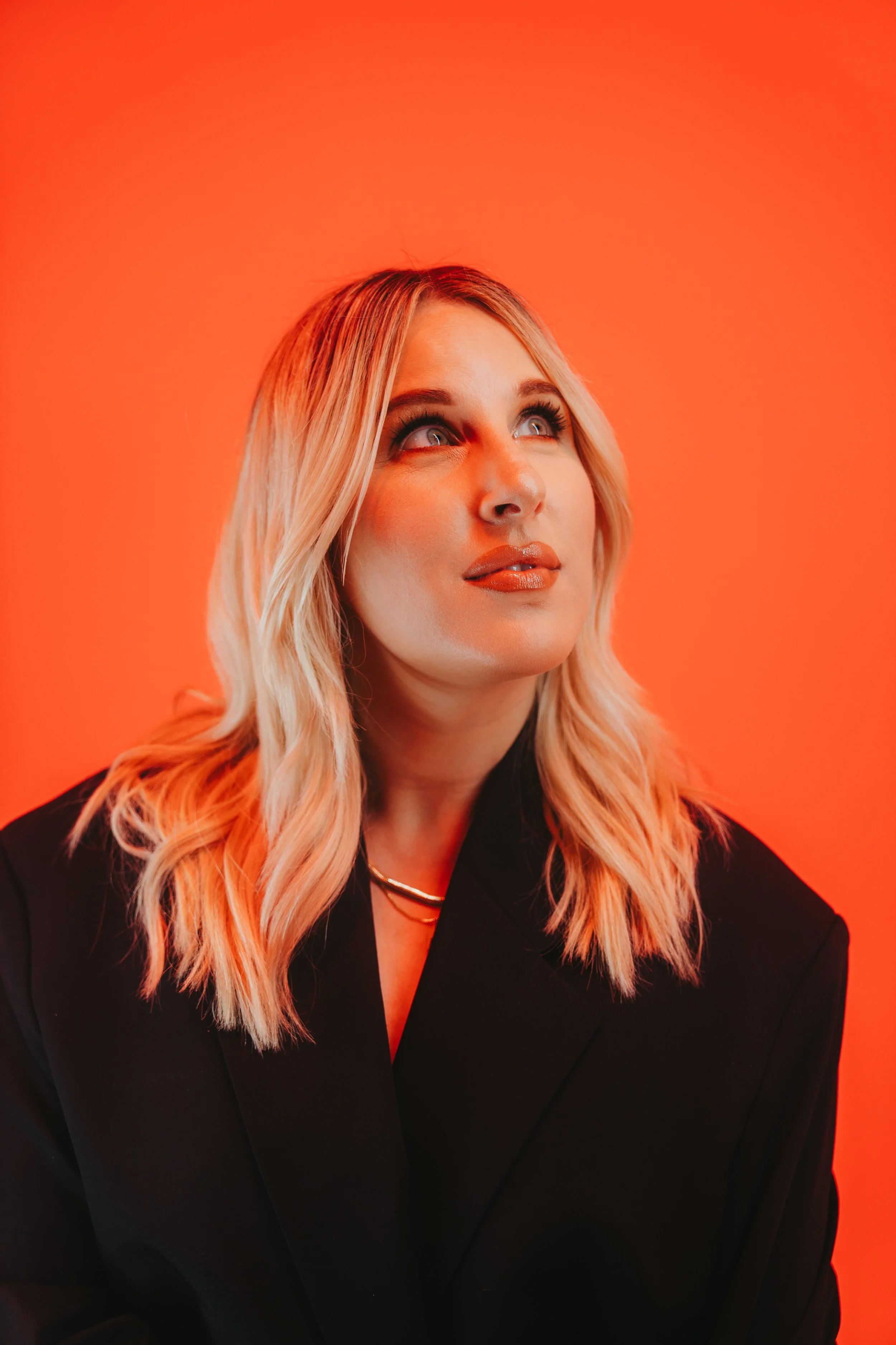 A woman with blonde hair and makeup looking upward against a bright orange background.
