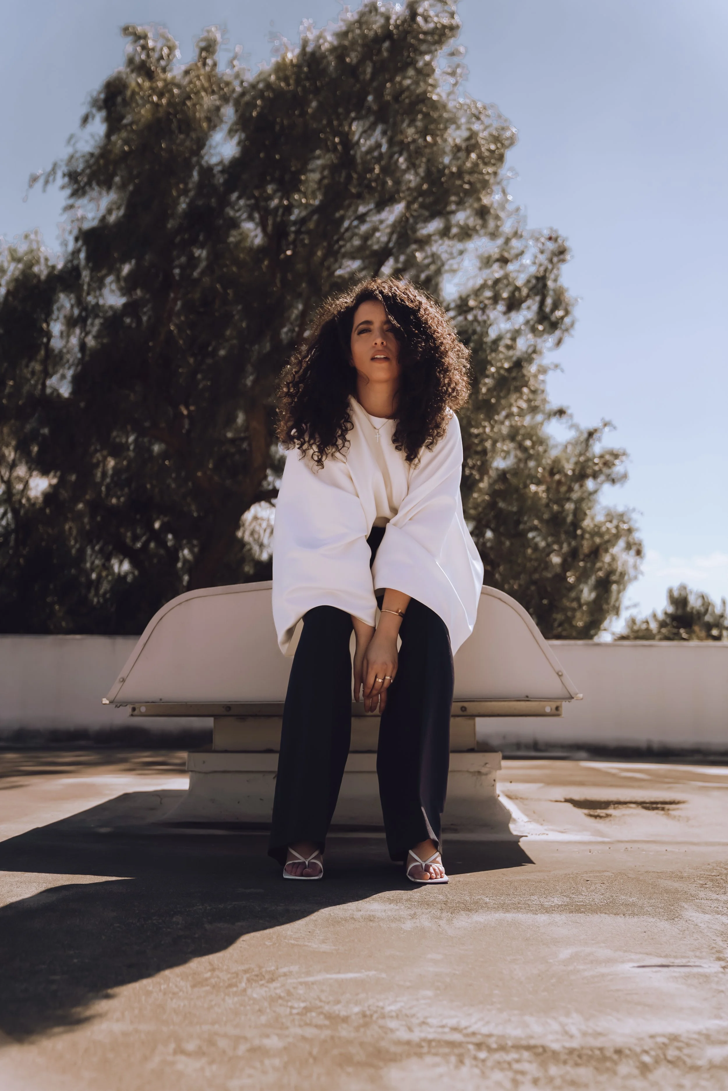A woman with curly hair sitting on a white bench outdoors, wearing a white top, black pants, and high heels, under a clear blue sky with large trees in the background.