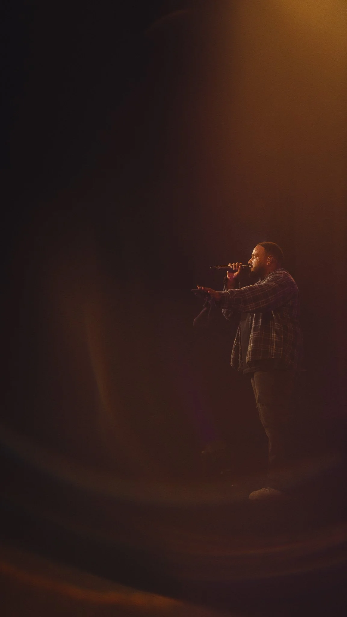 A person singing into a microphone on stage, illuminated by warm stage lighting.